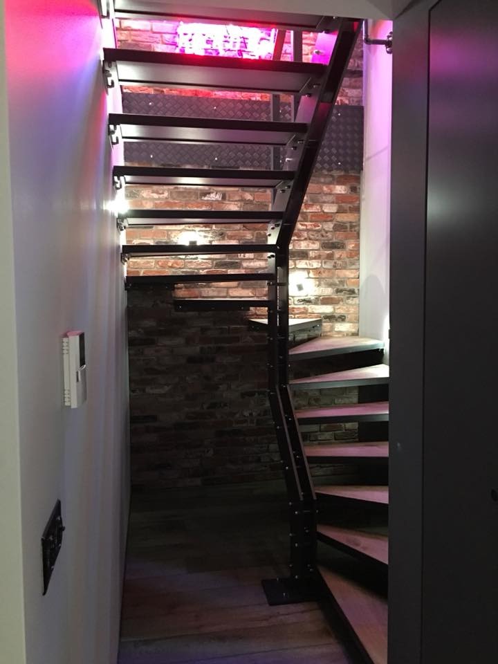 Nowoczesne schody metalowe z drewnianymi stopniami, oświetlone neonem i lampami ściennymi, na tle ściany z cegły i szarej szafy. Unikalny design w wąskim korytarzu.
