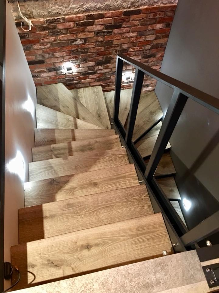 Drewniane schody z czarną balustradą, widok z góry. Czerwona cegła na ścianie i oświetlenie punktowe. Nowoczesny design z elementami industrialnymi.