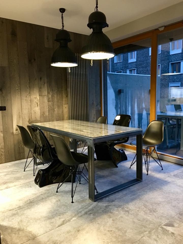 Stylowa jadalnia z industrialnymi lampami wiszącymi nad stołem z metalową ramą i szklanym blatem. Czarne krzesła o nowoczesnym designie dopełniają aranżację.