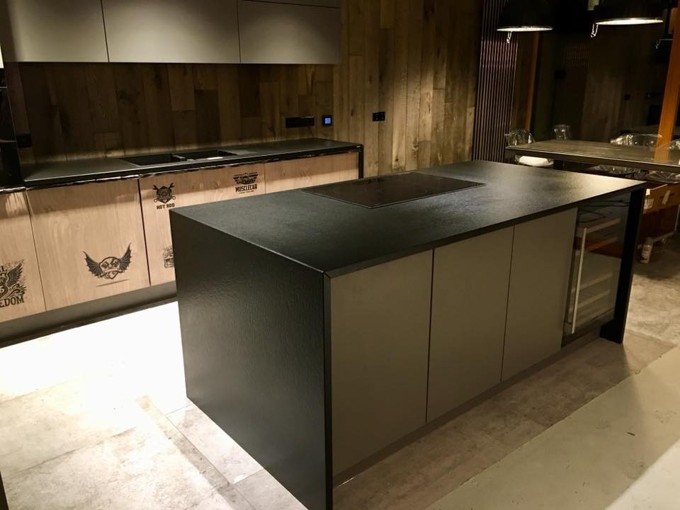 Nowoczesna kuchnia z wyspą i płytą indukcyjną. Fronty szafek zdobią motywy motoryzacyjne. Drewniane panele na ścianie dodają wnętrzu charakteru.