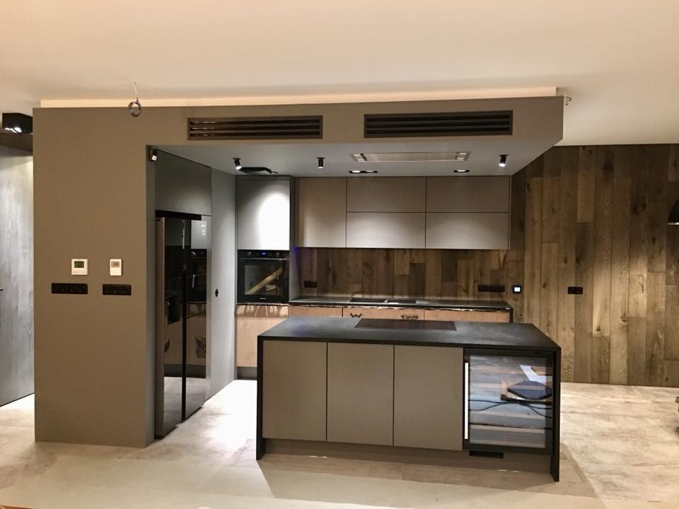 Nowoczesna kuchnia z wyspą i drewnianą ścianą. Minimalistyczne szafki, czarny blat, oświetlenie punktowe. Widoczne gniazdka elektryczne na ścianie.