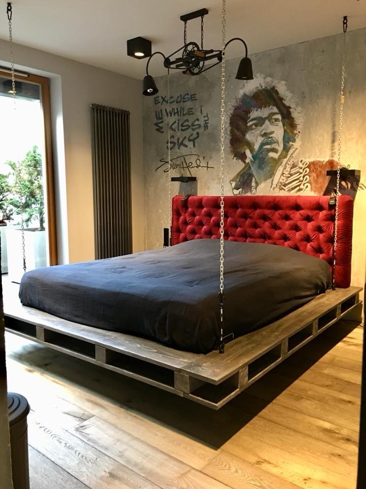 Nowoczesna sypialnia z wiszącym łóżkiem na łańcuchach, czerwonym zagłówkiem i industrialną lampą sufitową. Na ścianie mural z wizerunkiem muzyka.