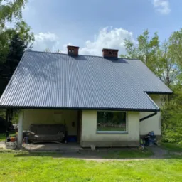 Dom z nowym, ciemnoszarym dachem z blachy trapezowej, widoczne dwa kominy z cegły, rynna w kolorze dachu, trawnik z mleczami na pierwszym planie.