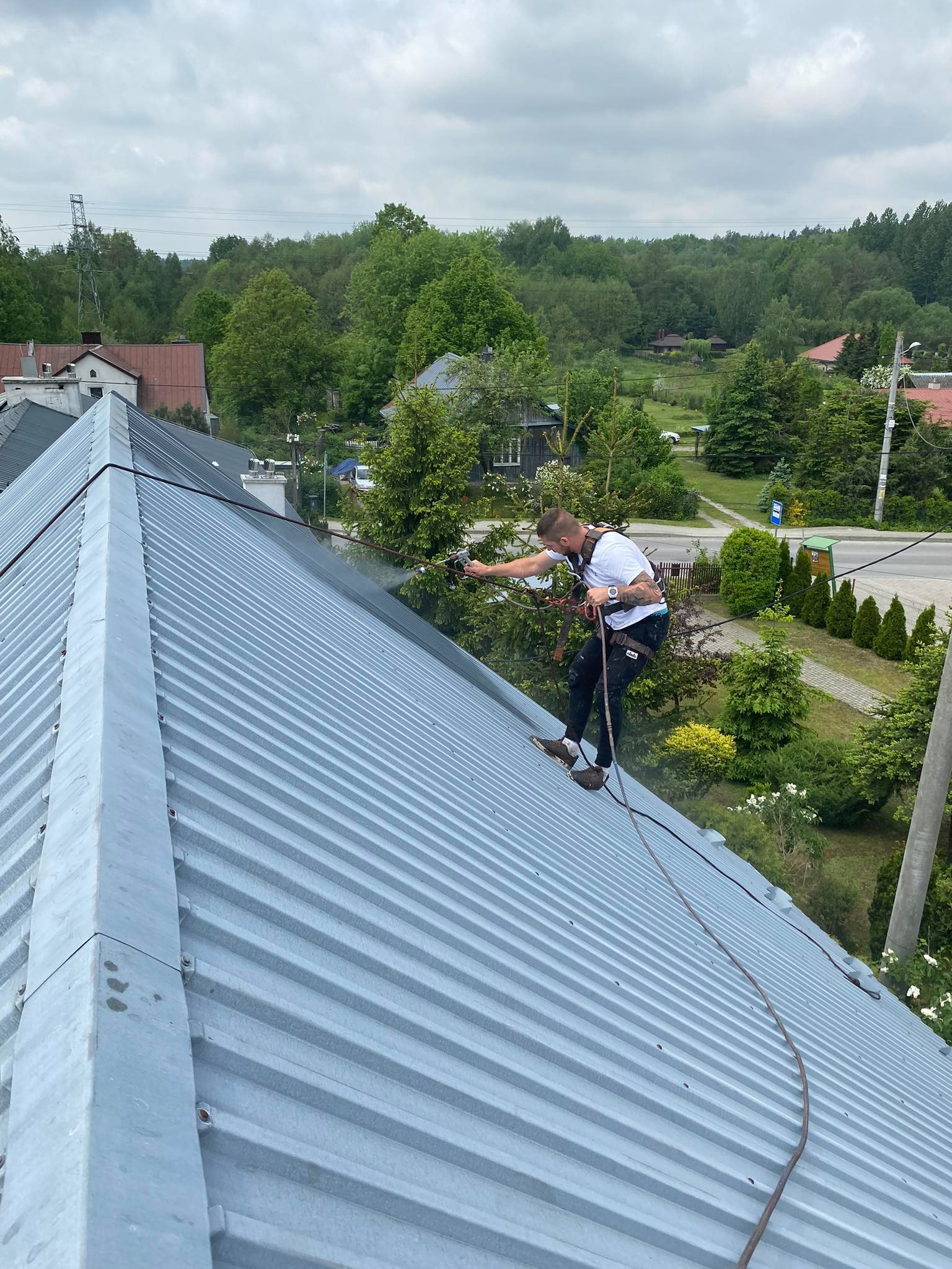 Pracownik w uprzęży zabezpieczającej myje metalowy dach z użyciem lancy ciśnieniowej; widoczny efekt czyszczenia, zieleń w tle.