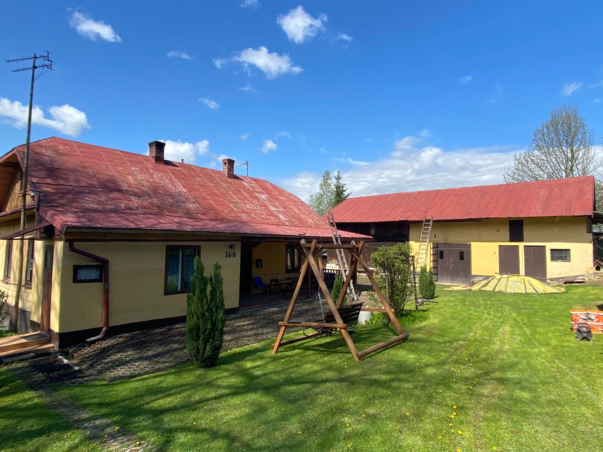Dwupiętrowy dom z czerwonym dachem i przylegający budynek gospodarczy, również z czerwonym dachem, widziane z perspektywy ogrodu z huśtawką i rozłożoną drabiną opartą o dach budynku gospodarczego.