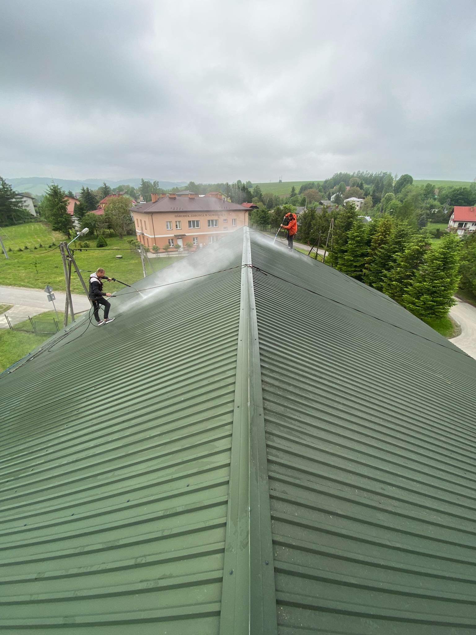 Dwóch mężczyzn czyści zielony dach z blachy falistej za pomocą myjek ciśnieniowych, w tle budynek i zieleń.