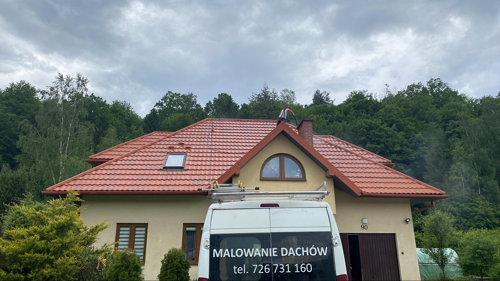 Pracownik myje ciśnieniowo dach domu jednorodzinnego z czerwonej dachówki; na pierwszym planie samochód dostawczy z napisem 'Malowanie Dachów' i numerem telefonu.