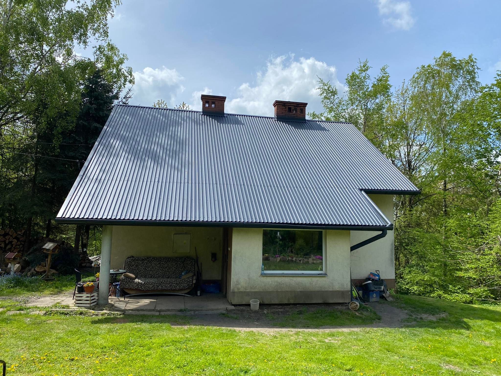 Dom z nowym, ciemnoszarym dachem z blachy trapezowej, widoczne dwa kominy z cegły, rynna w kolorze dachu, trawnik z mleczami na pierwszym planie.
