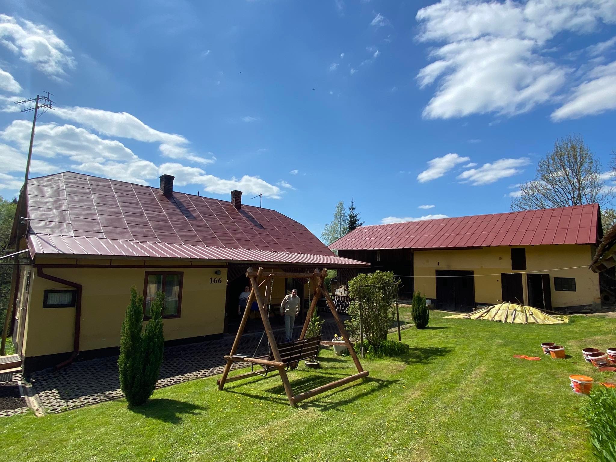 Żółty dom z nowym, czerwonym dachem blachodachówkowym, widoczny z perspektywy ogrodu z huśtawką i pojemnikami po farbie.