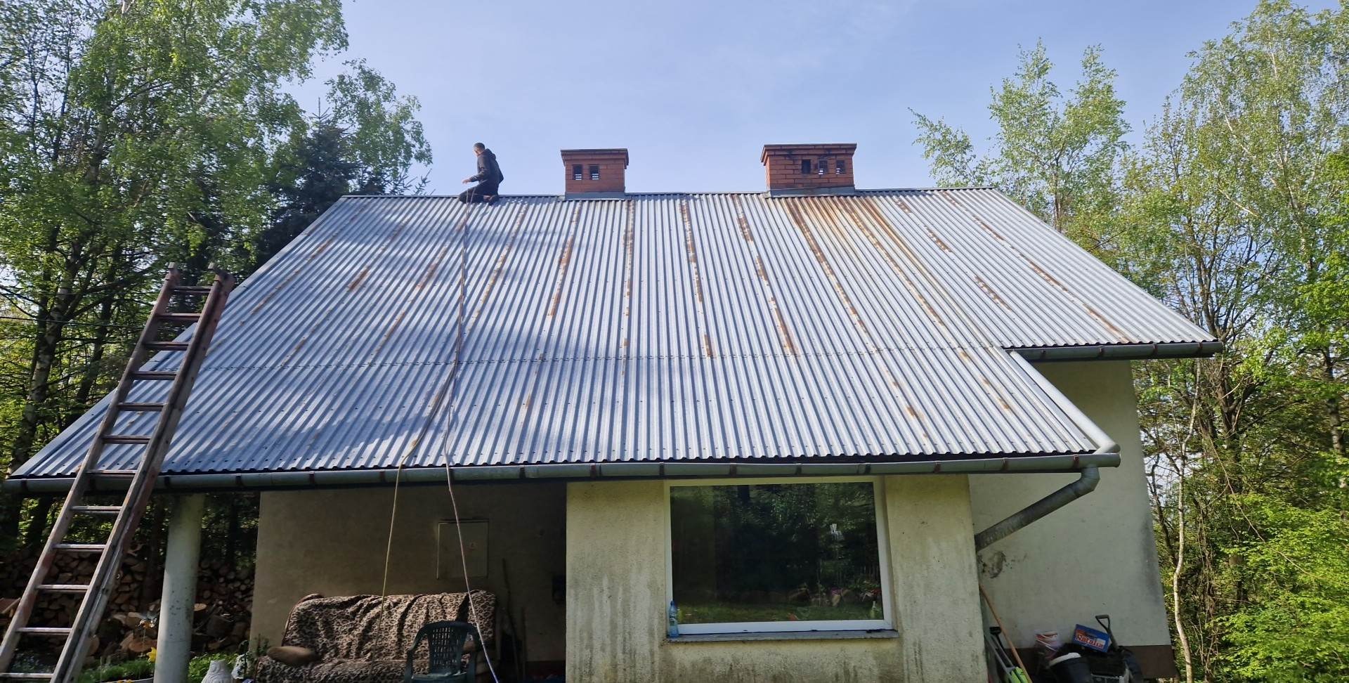 Pracownik siedzi na blaszanej, pofałdowanej powierzchni dachu domu jednorodzinnego, obok dwóch ceglanych kominów. Do ściany oparta drabina. Widoczne zacieki na elewacji i rynna.