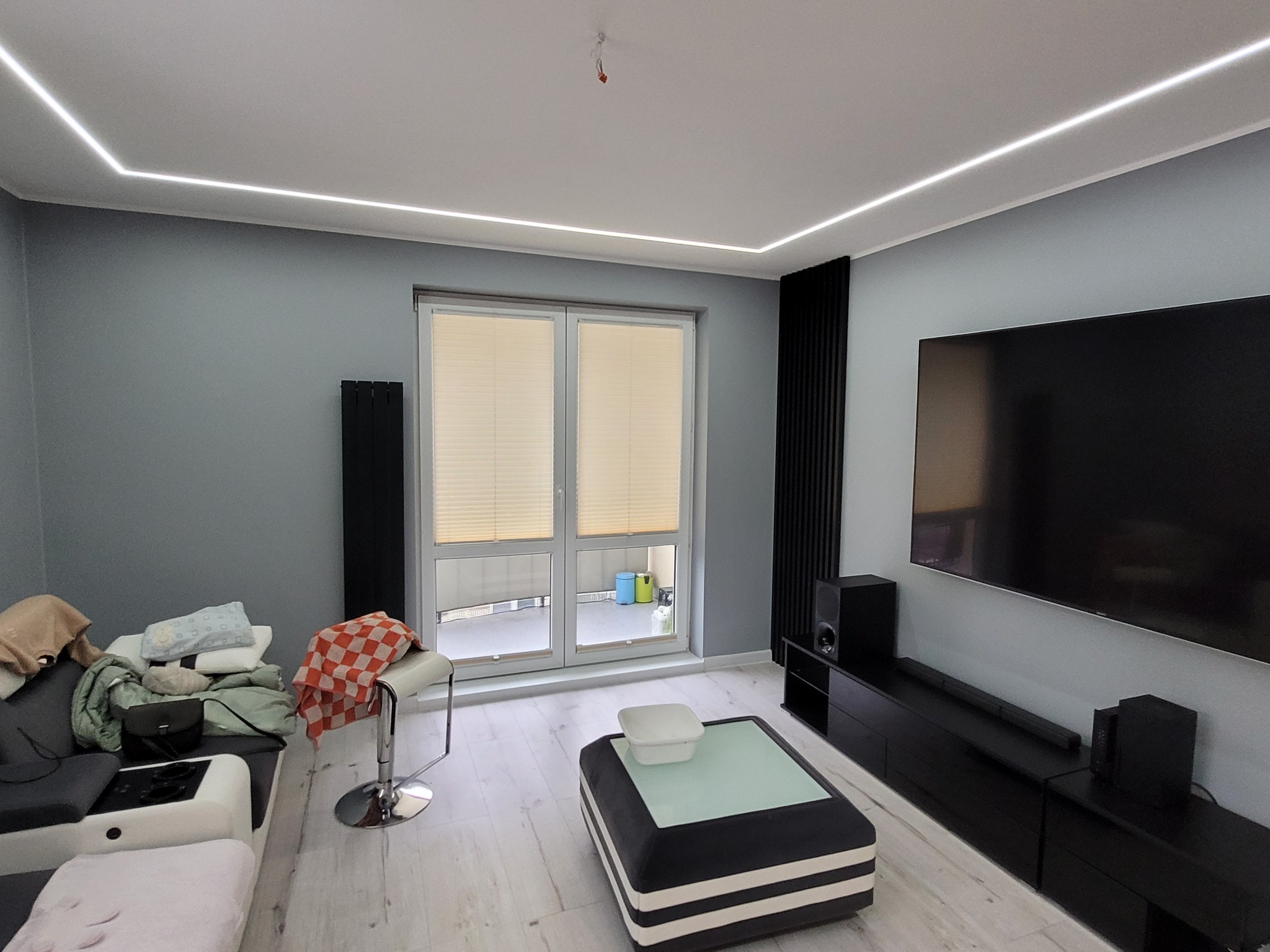 Szare wnętrze salonu z oświetleniem LED podsufitowym, czarnymi panelami dekoracyjnymi na ścianie, telewizorem i meblami w stylu nowoczesnym.