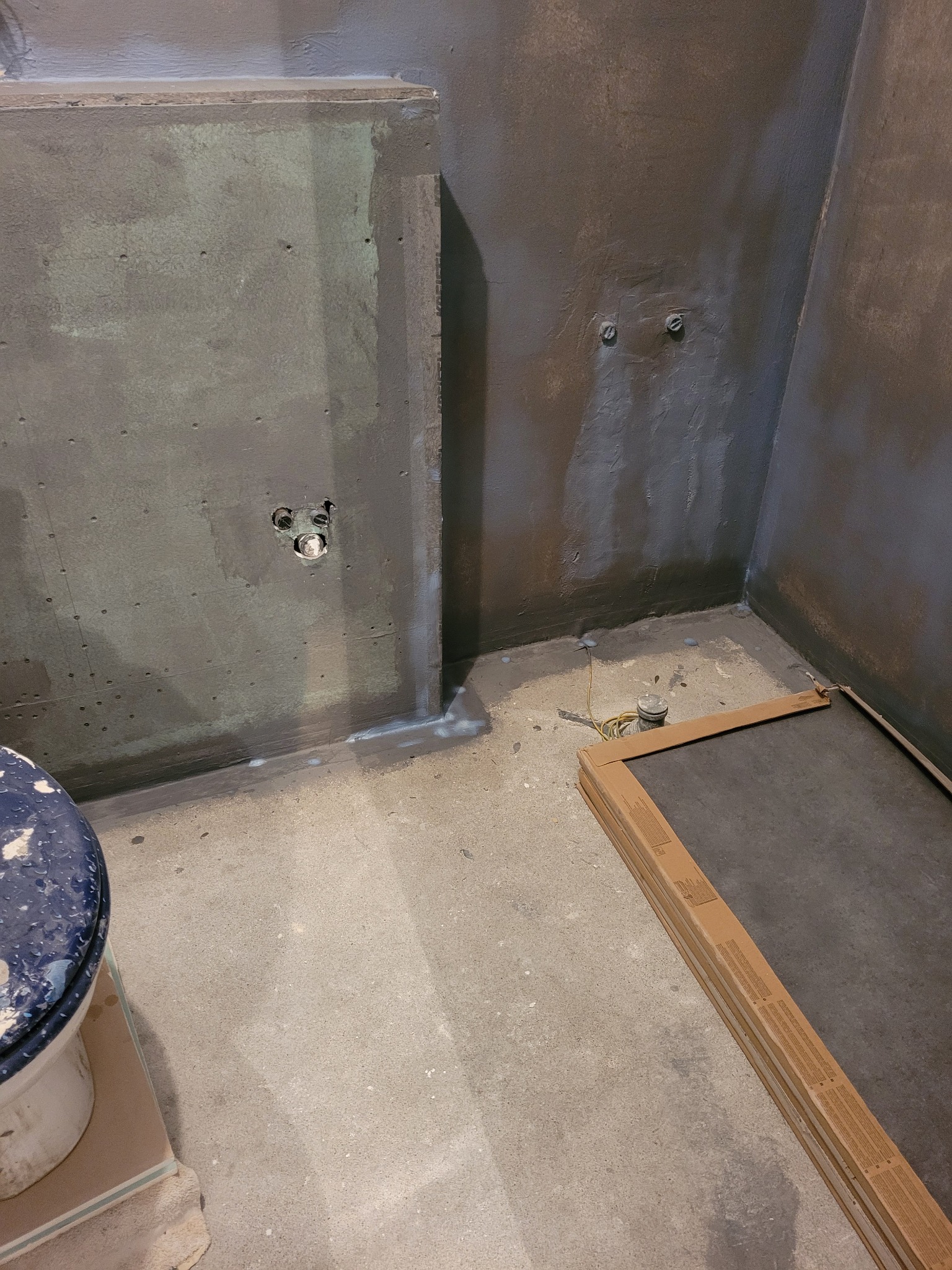 Przygotowana do wykończenia łazienka z uszczelnionymi ścianami, widoczne puszki elektryczne i fragment toalety, na podłodze ułożone płyty.