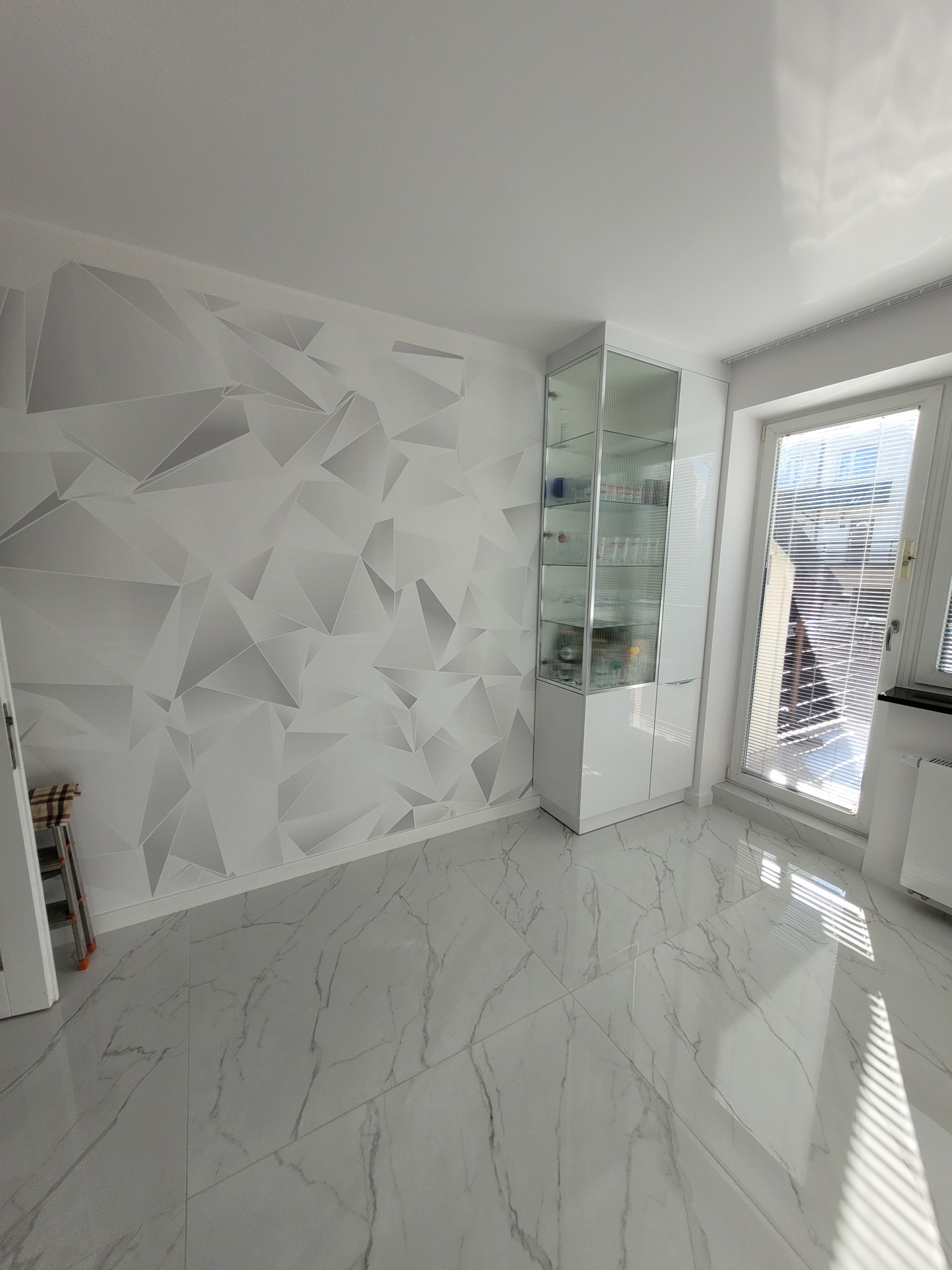 Jasne wnętrze po remoncie: białe marmurowe płytki podłogowe, ściana z geometrycznym wzorem w odcieniach szarości, biała witryna z przeszkleniami i okno z żaluzjami.