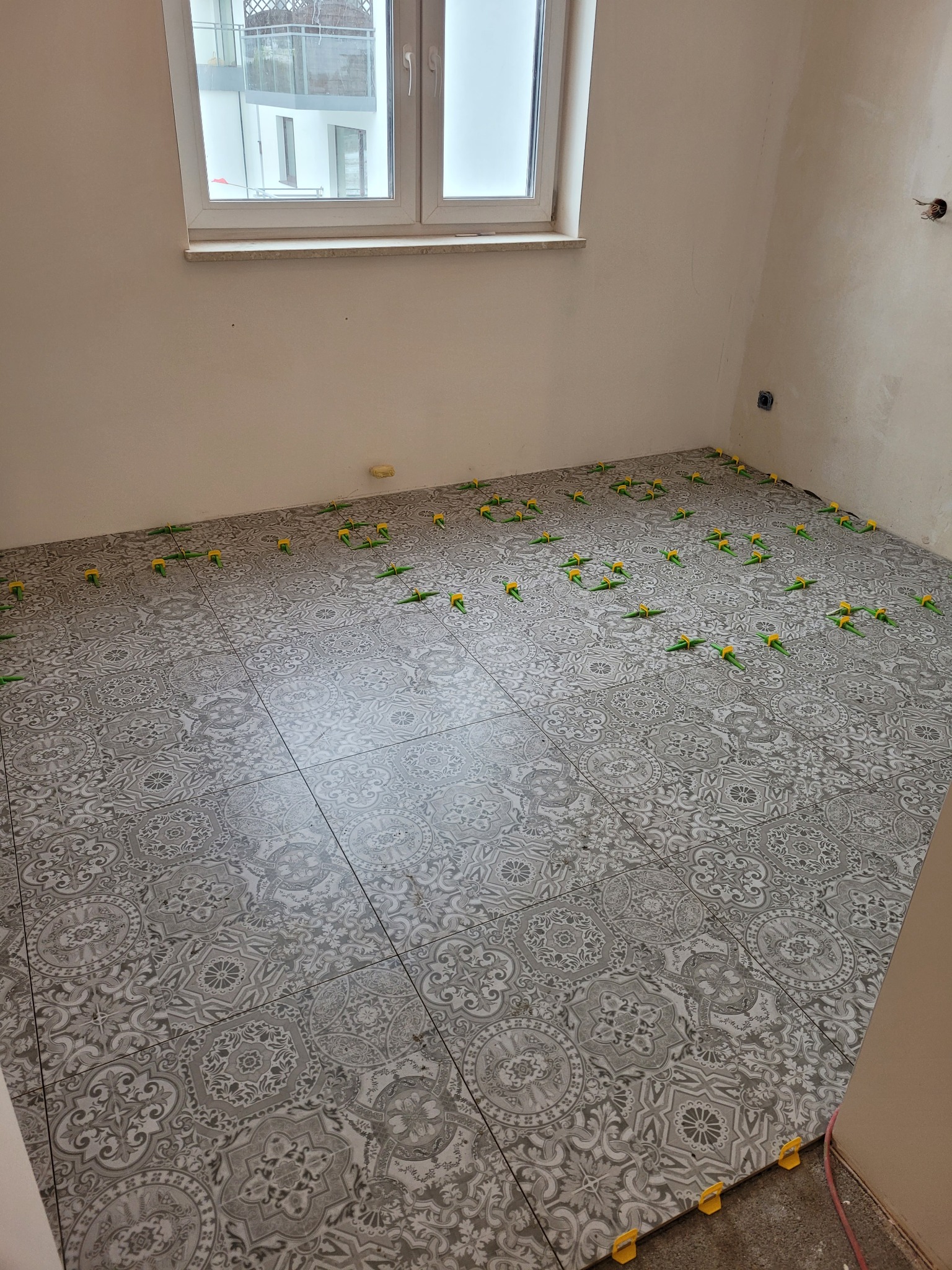 Szare płytki podłogowe z dekoracyjnym wzorem ułożone w pokoju, z widocznymi zielono-żółtymi klipsami poziomującymi na świeżo położonej podłodze, przy ścianie widoczne gniazdko elektryczne.