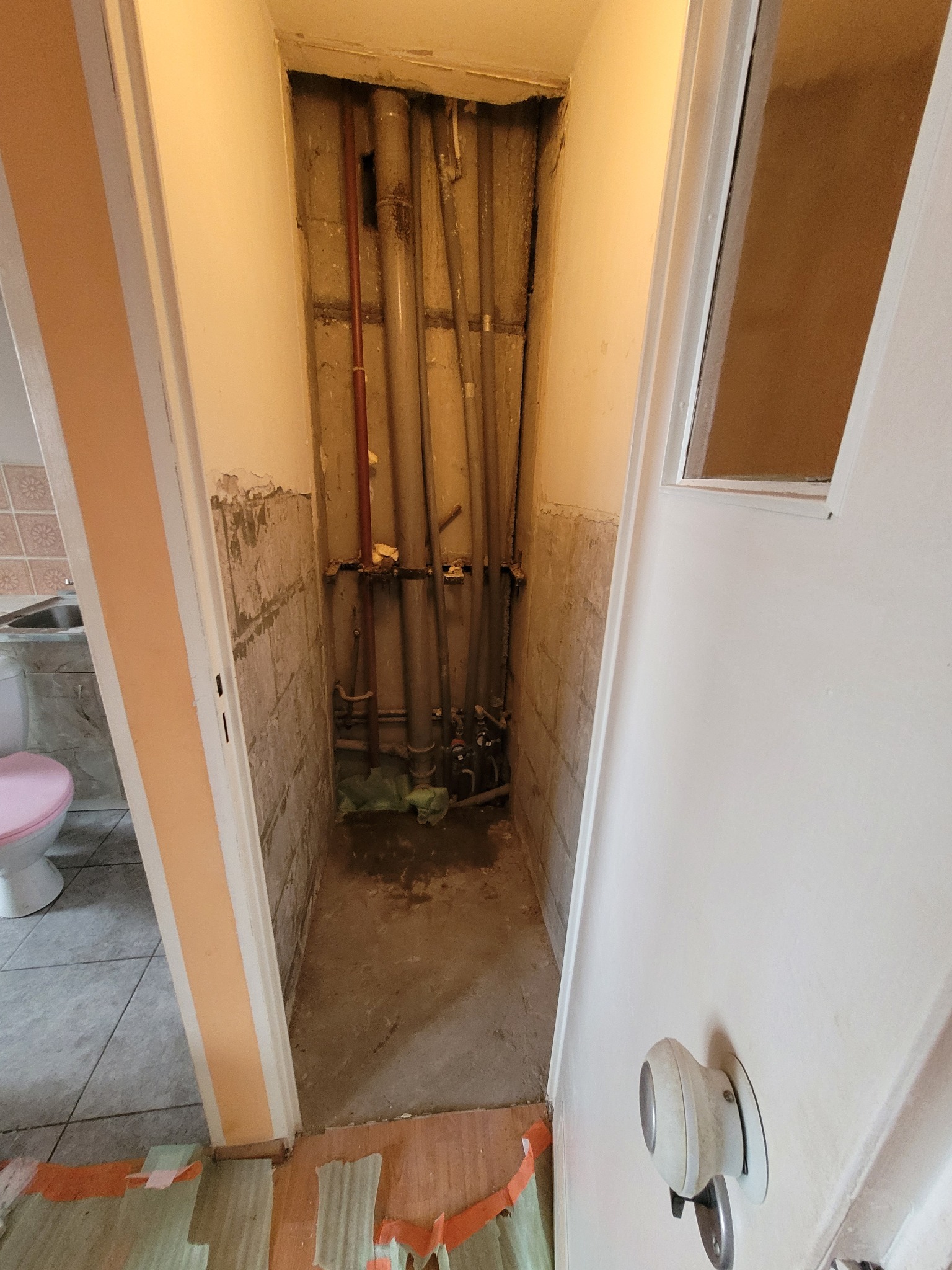 Wnęka w łazience w trakcie prac remontowych: odsłonięte rury, częściowo usunięte płytki, widoczne ślady po skuciu, zabezpieczona podłoga, widok na toaletę i umywalkę.