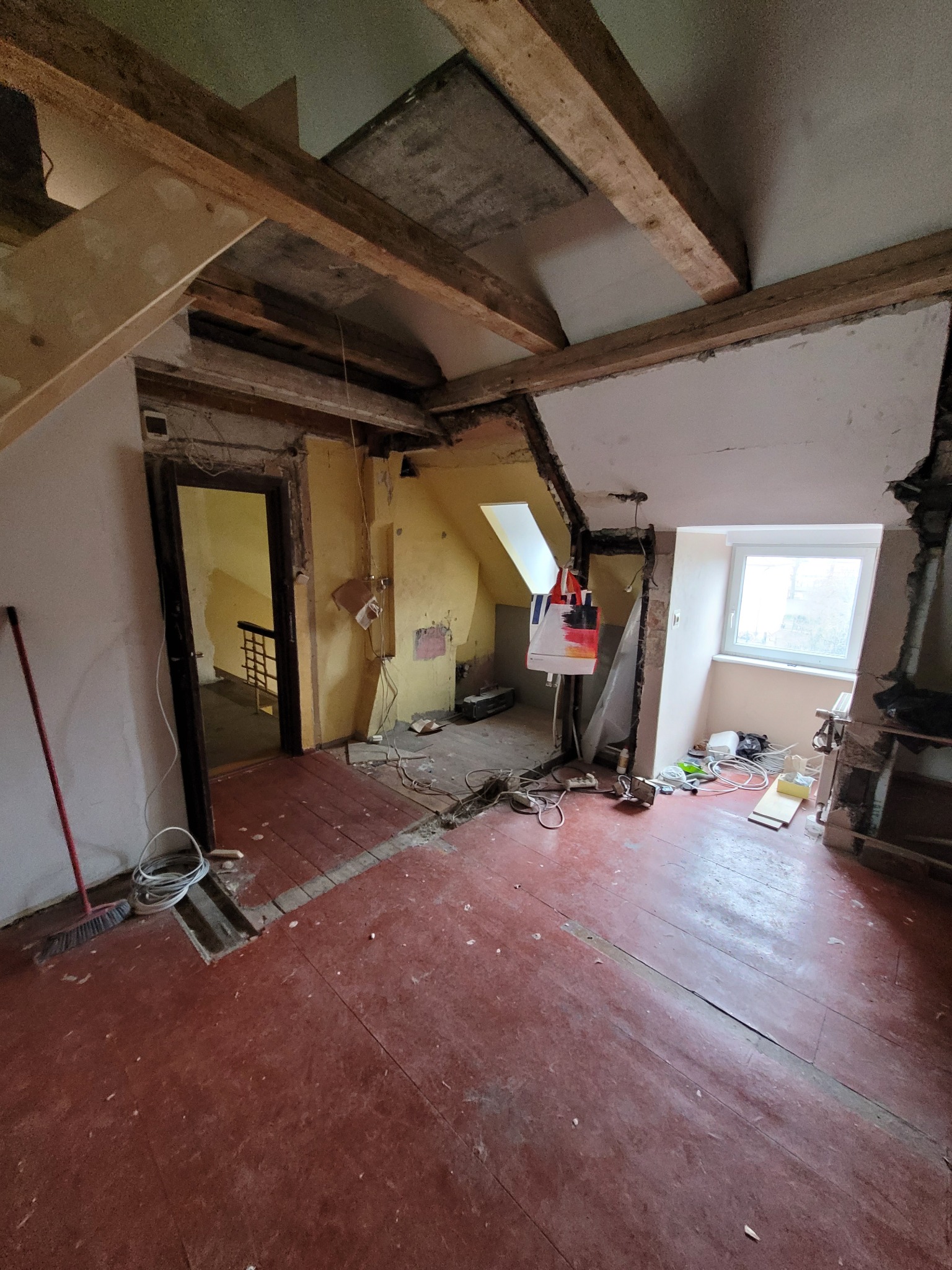 Pokój na poddaszu w trakcie remontu: odsłonięte belki stropowe, częściowo otynkowane ściany, stara, czerwona podłoga z desek, widoczne kable i narzędzia, okno z widokiem na zewnątrz.