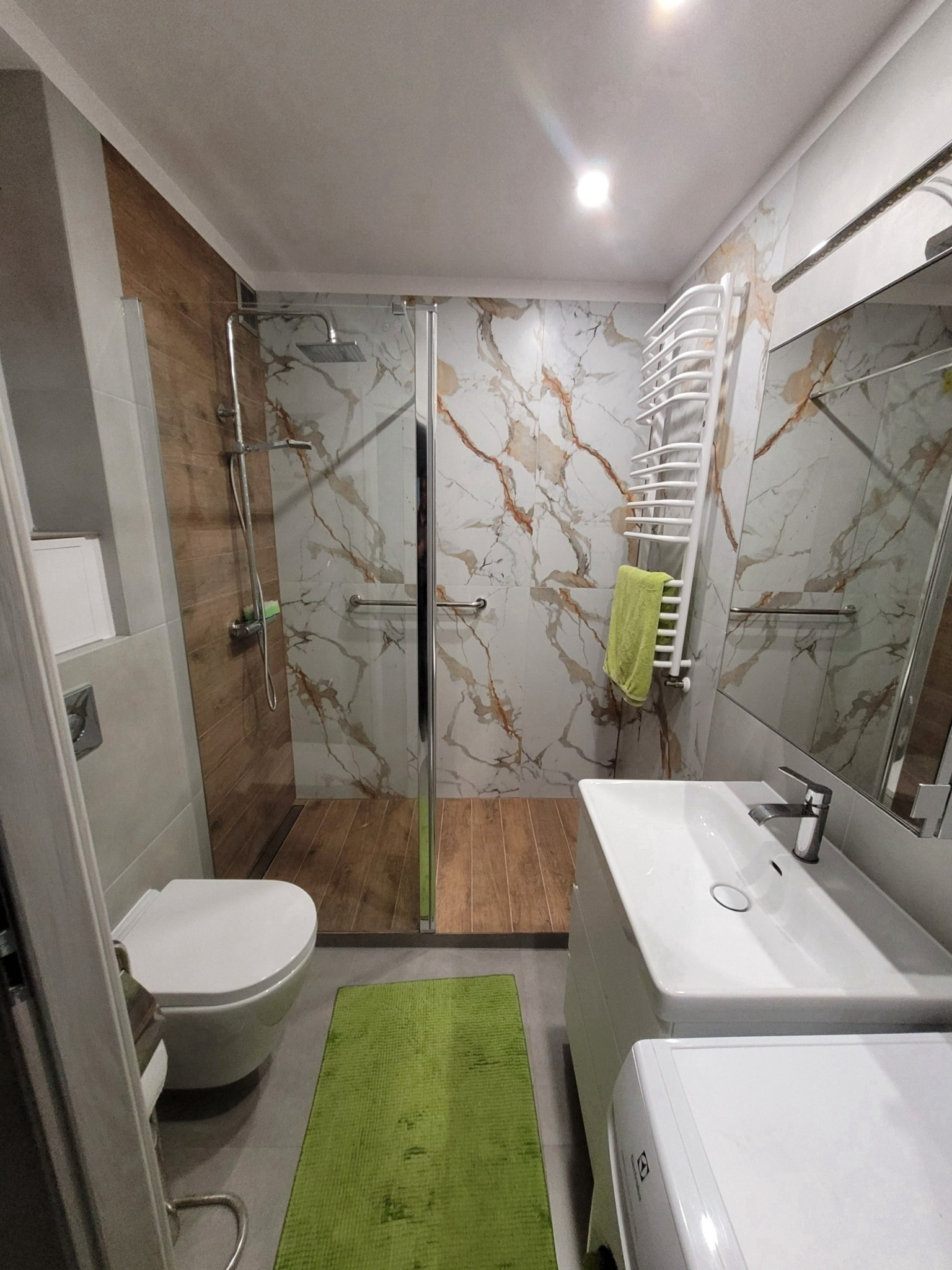 Nowoczesna łazienka z kabiną prysznicową typu walk-in, wykończona płytkami imitującymi marmur i drewno, z zielonym ręcznikiem na grzejniku i zielonym dywanikiem.