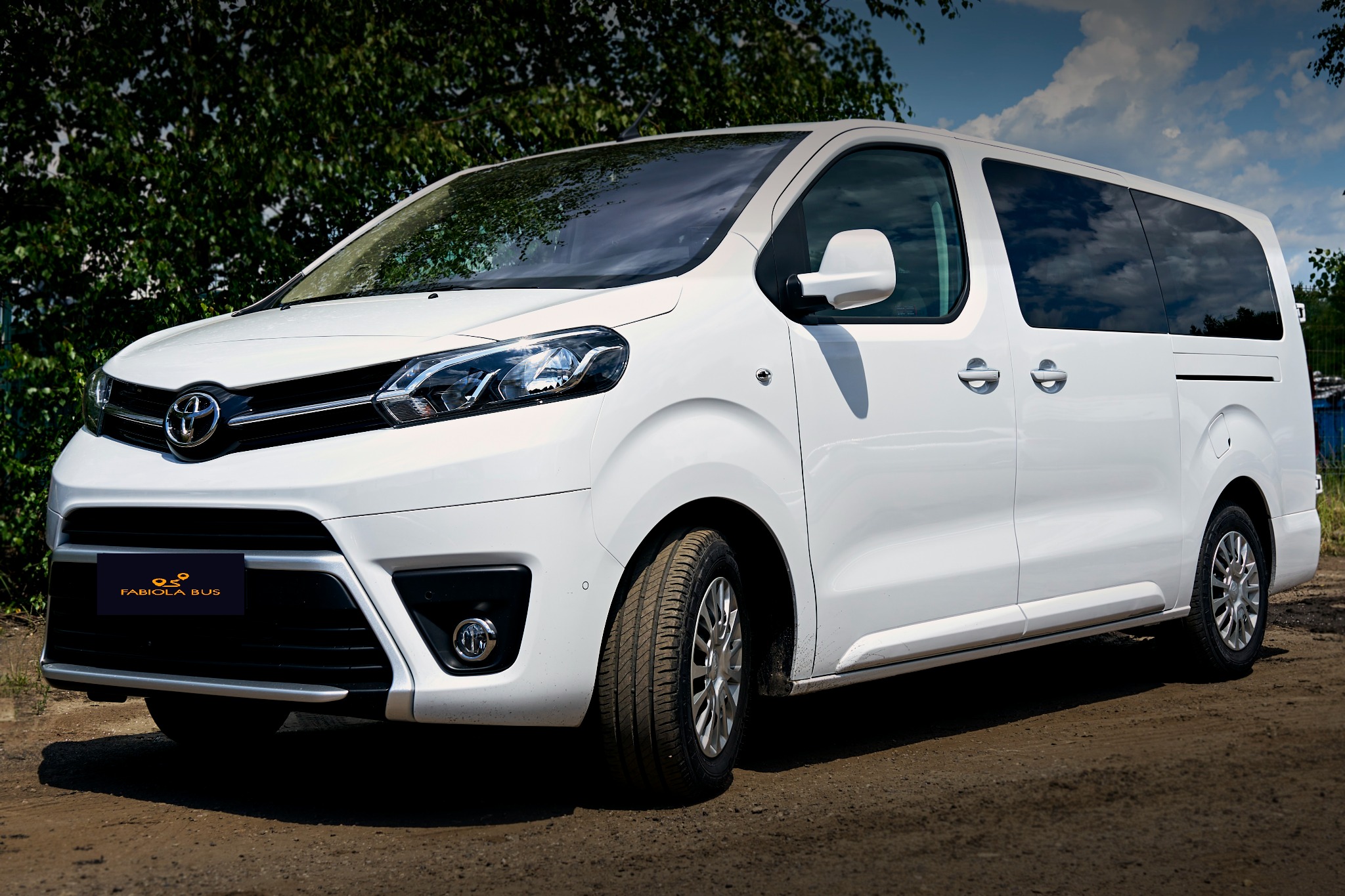 Biały bus Toyota Proace Verso z logo Fabiola Bus na tle zieleni i błękitnego nieba.