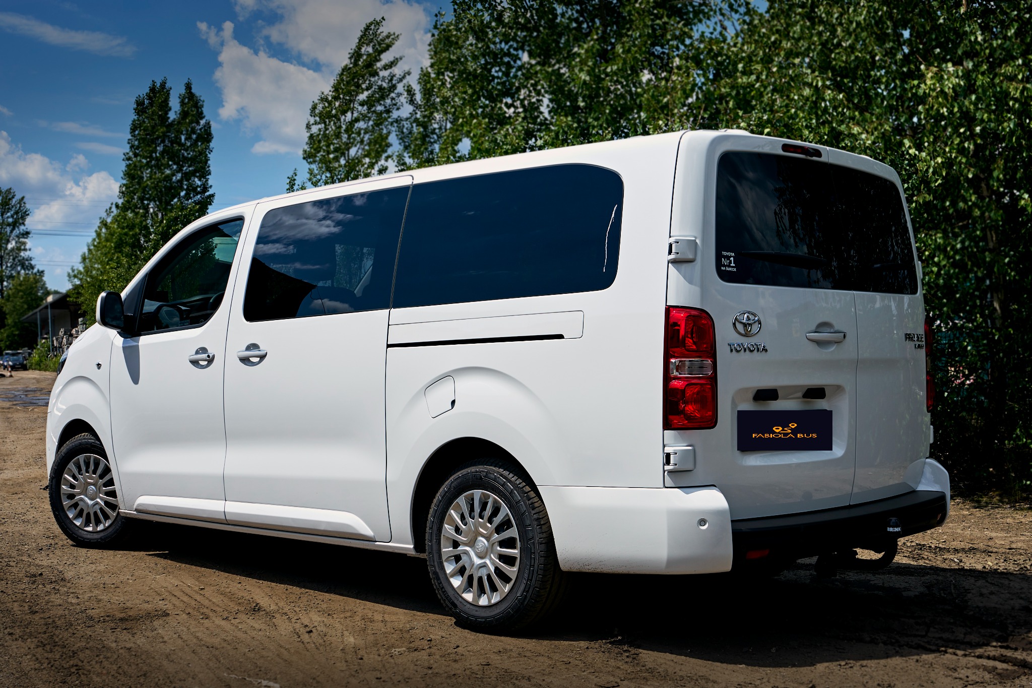 Biały bus Toyota Proace Long z przyciemnianymi szybami, widok z tyłu na tle drzew i nieba, z naklejką 'Toyota Nr. 1 na świecie' na tylnej szybie i logo 'Fabiola Bus' na klapie bagażnika.