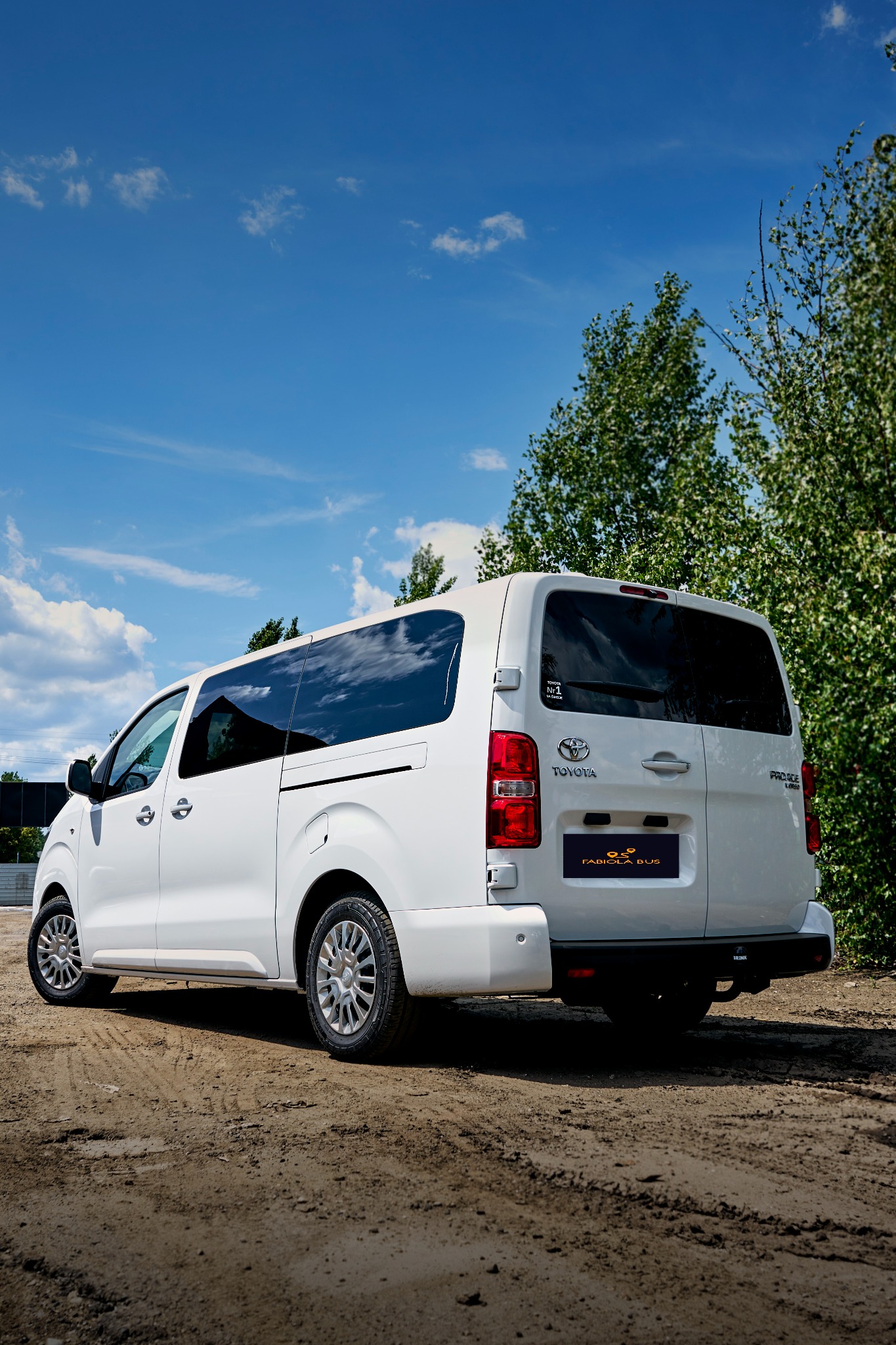 Biały bus Toyota Proace Verso z przyciemnianymi szybami, widok z tyłu na tle zieleni i błękitnego nieba z chmurami, logo firmy Fabiola Bus na tylnych drzwiach.