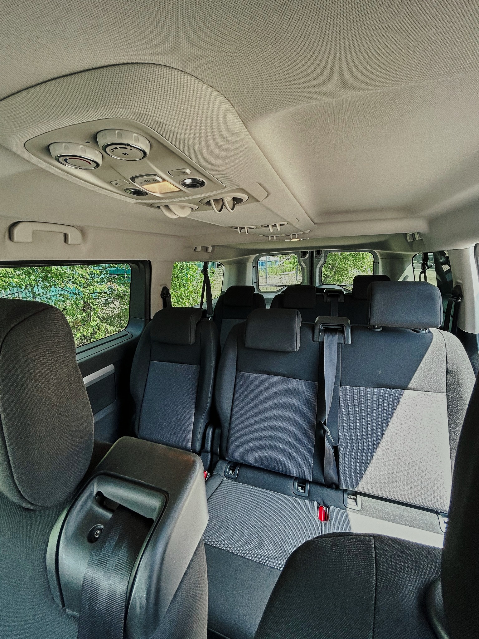 Wnętrze przestronnego busa z trzema rzędami siedzeń, widok z tyłu, z zielenią za oknami i regulacją klimatyzacji na suficie.