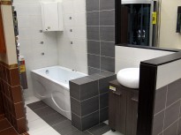 Nowoczesna łazienka z białą wanną z hydromasażem, umywalką nablatową i szaro-białymi płytkami. Widoczna zabudowa wanny z szarych płytek oraz szafka pod umywalką w kolorze ciemnego drewna.