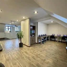 Przestronne wnętrze sali komputerowej z drewnianą podłogą, rzędem biurek z laptopami i krzesłami, przeszklonym pomieszczeniem z oknami w tle oraz rośliną doniczkową, oświetlone punktowymi lampami...