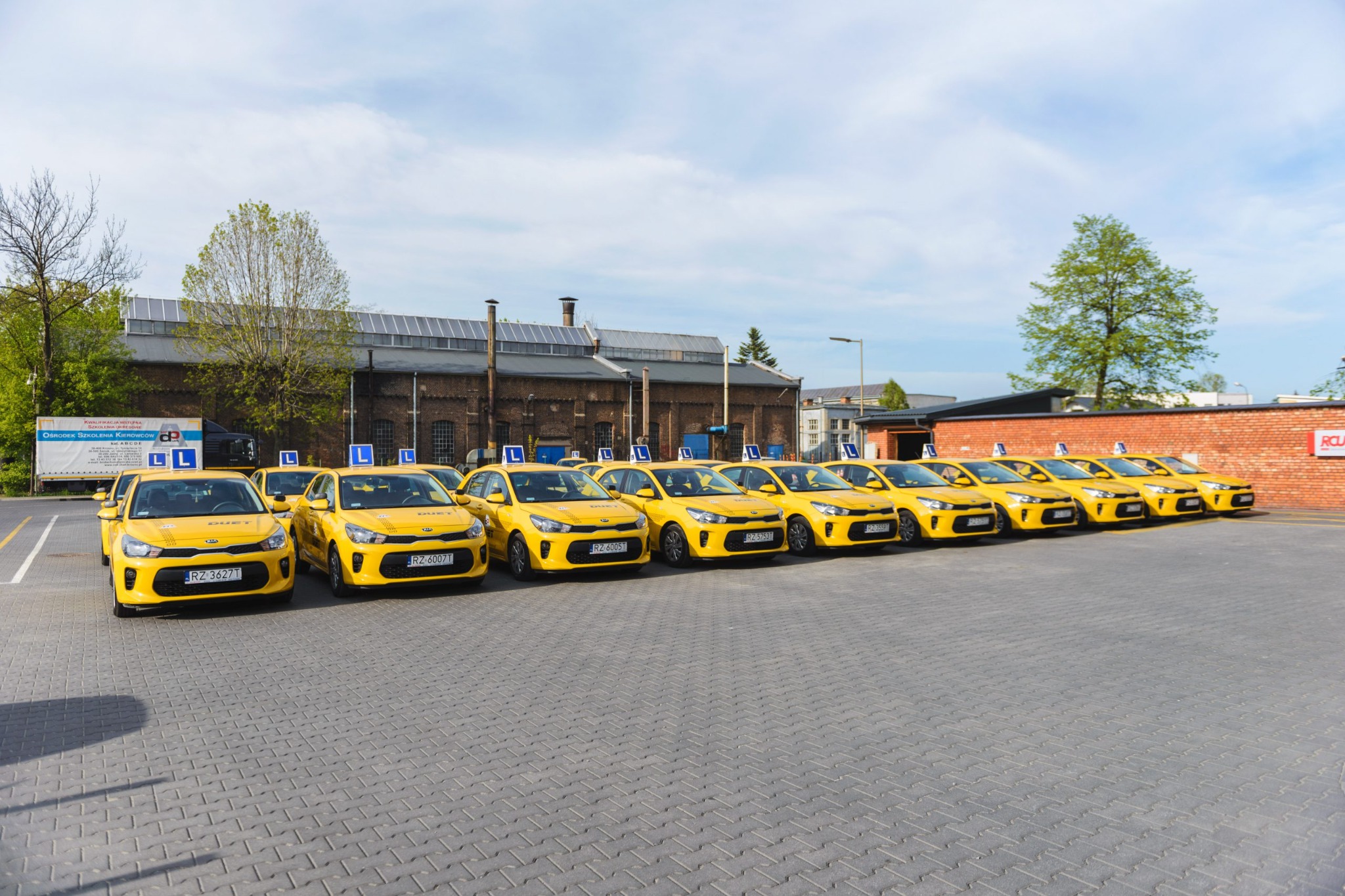 Rząd żółtych samochodów z oznaczeniem 'L' na dachu, zaparkowanych na brukowanym placu przed budynkiem, prawdopodobnie flota pojazdów do nauki jazdy.