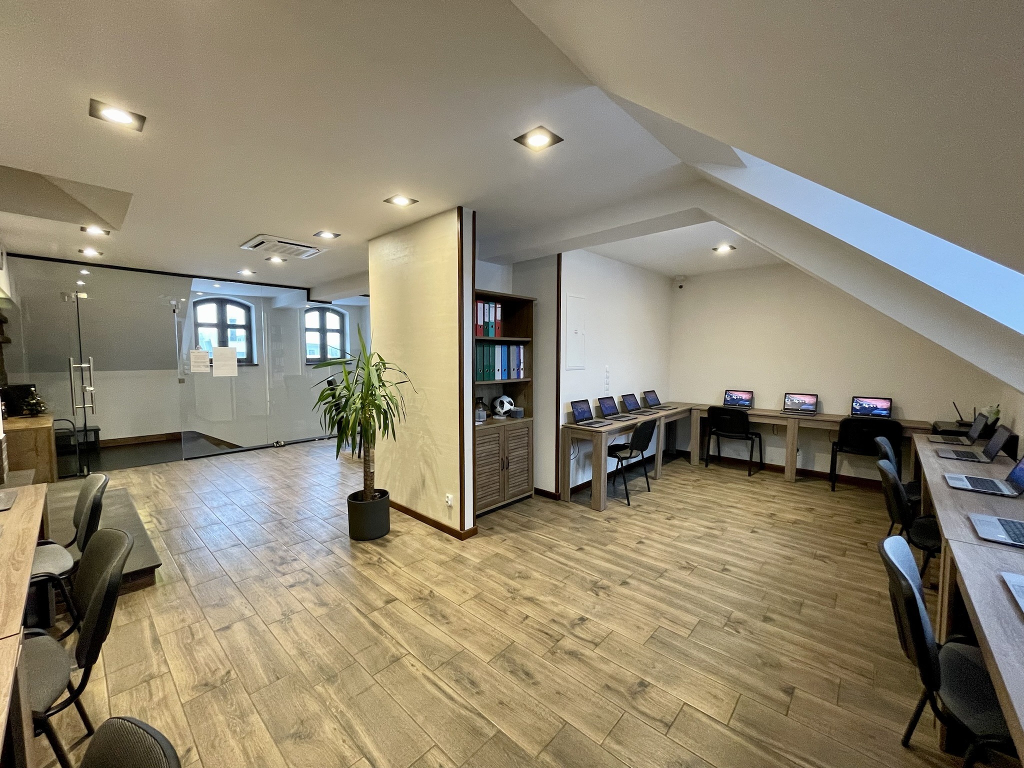 Przestronne wnętrze sali komputerowej z drewnianą podłogą, rzędem biurek z laptopami i krzesłami, przeszklonym pomieszczeniem z oknami w tle oraz rośliną doniczkową, oświetlone punktowymi lampami...