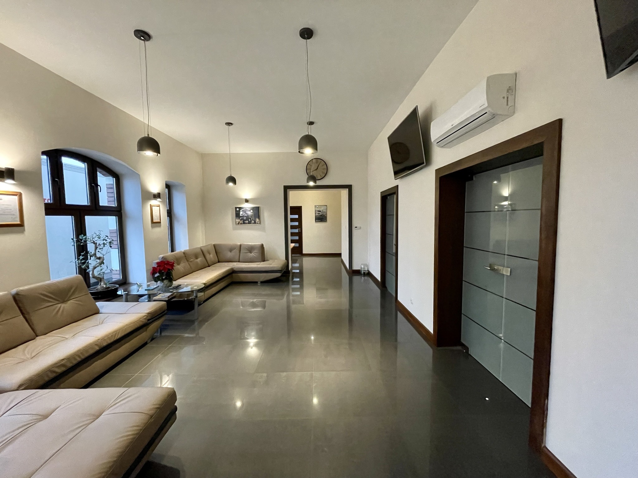 Przestronne, nowocześnie urządzone wnętrze poczekalni z beżowymi sofami narożnymi, szarymi płytkami podłogowymi z połyskiem, wiszącymi lampami i klimatyzacją.