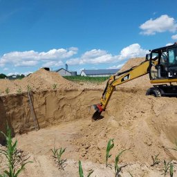 AUTO-ZAMIELA SKP Przemysław Zamiela - Żółta koparka CAT M315D w trakcie prac ziemnych, pogłębiania wykopu o okrągłym kształcie, widoczne ślady pomiarów na ziemi, w tle budynki gospodarcze i błękitne niebo z chmurami.