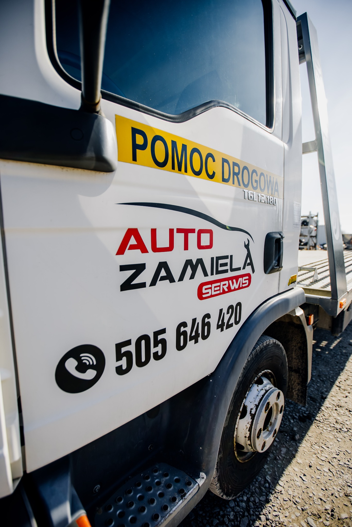 Biała laweta z żółtym napisem 'POMOC DROGOWA' i czerwonym 'AUTO ZAMIELA SERWIS' oraz numerem telefonu, stojąca na żwirowym podłożu.
