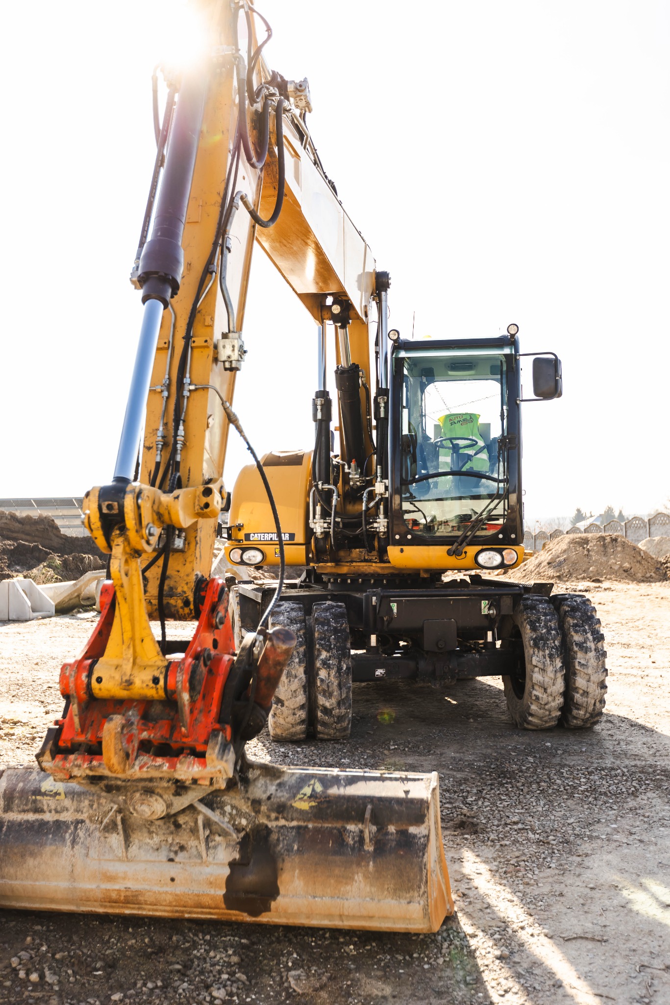 Żółto-czarna koparka kołowa Caterpillar z brudną łyżką i pomarańczowym szybkozłączem na żwirowym placu budowy, operator widoczny w kabinie, ostre światło słoneczne z góry.