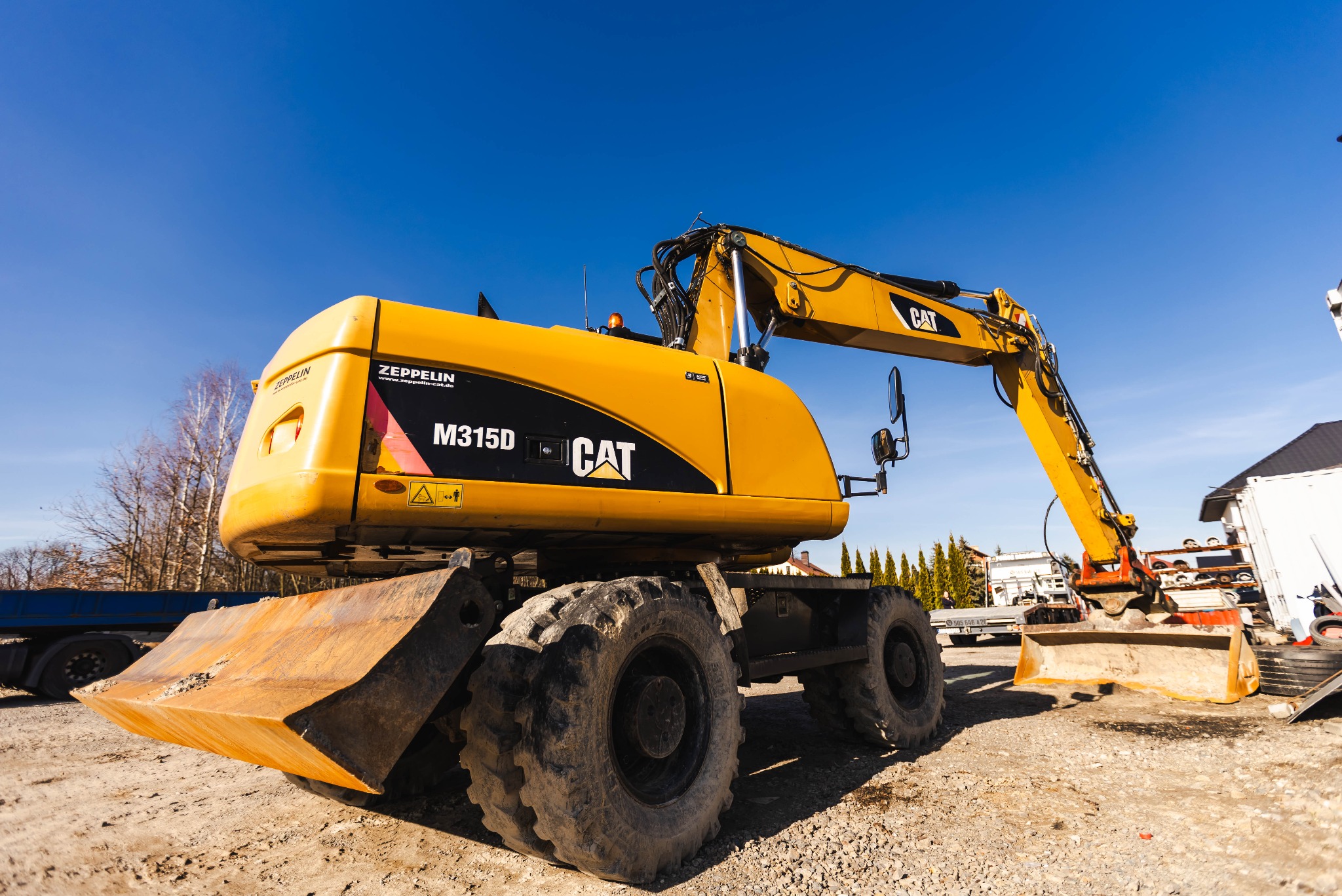 Żółta koparka kołowa CAT M315D na placu budowy, widok z dołu, błękitne niebo w tle.