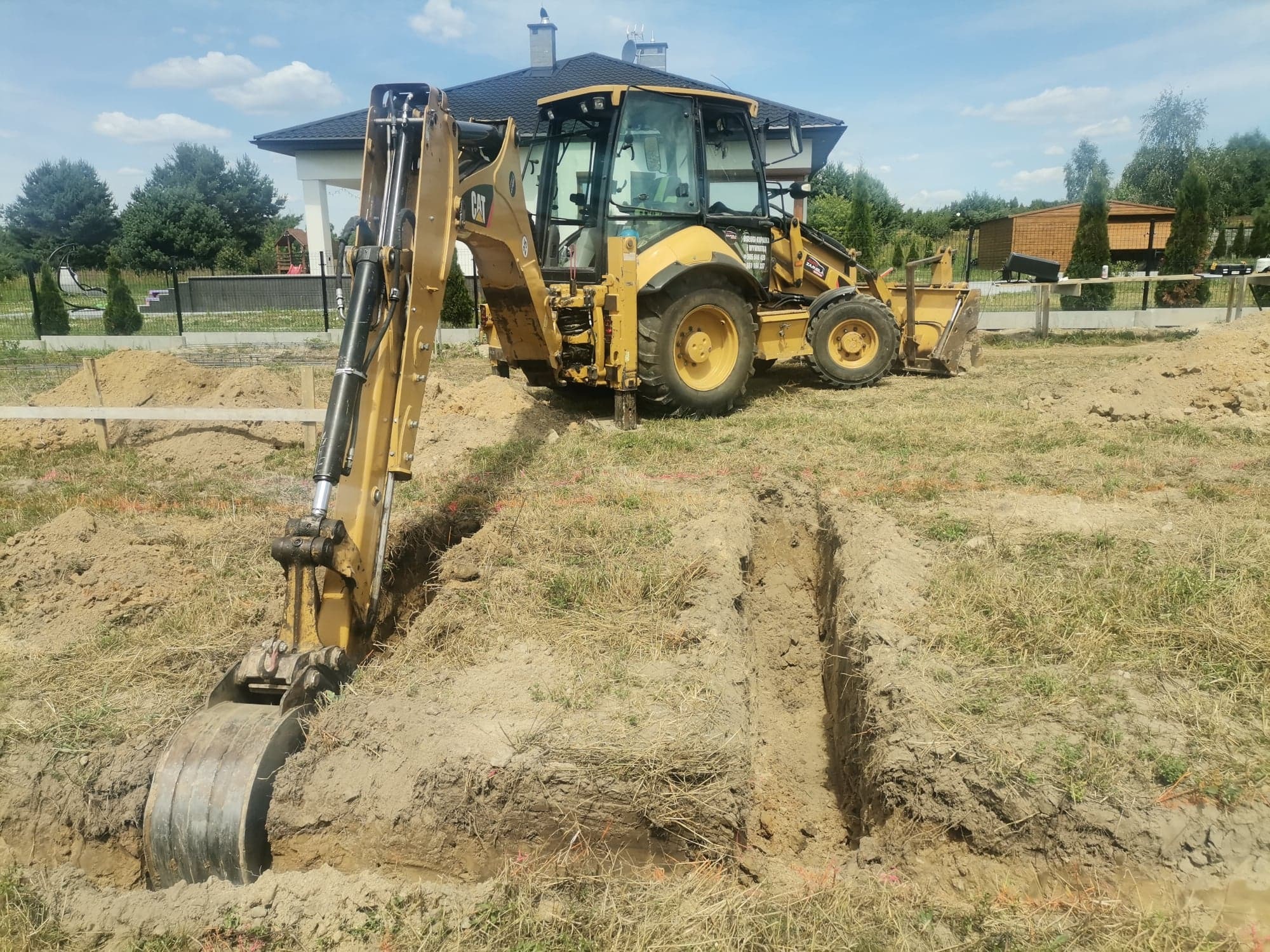 Żółta koparko-ładowarka CAT wykonuje wykop liniowy w piaszczystej ziemi na budowie domu jednorodzinnego z czarnym dachem.