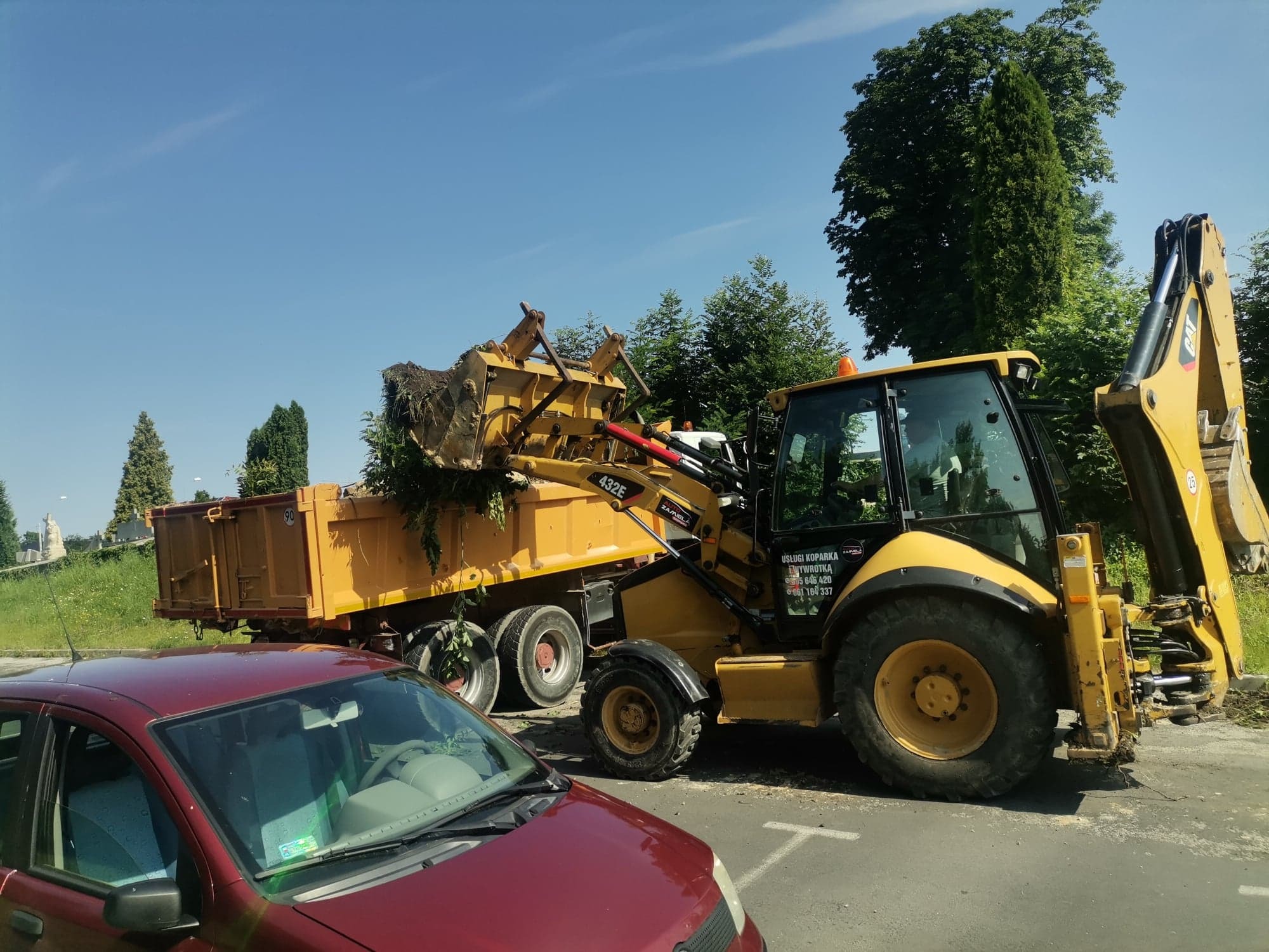 Żółta koparko-ładowarka załadowuje ziemię z zielenią do skrzyni ciężarówki; w tle drzewa i czerwony samochód osobowy.