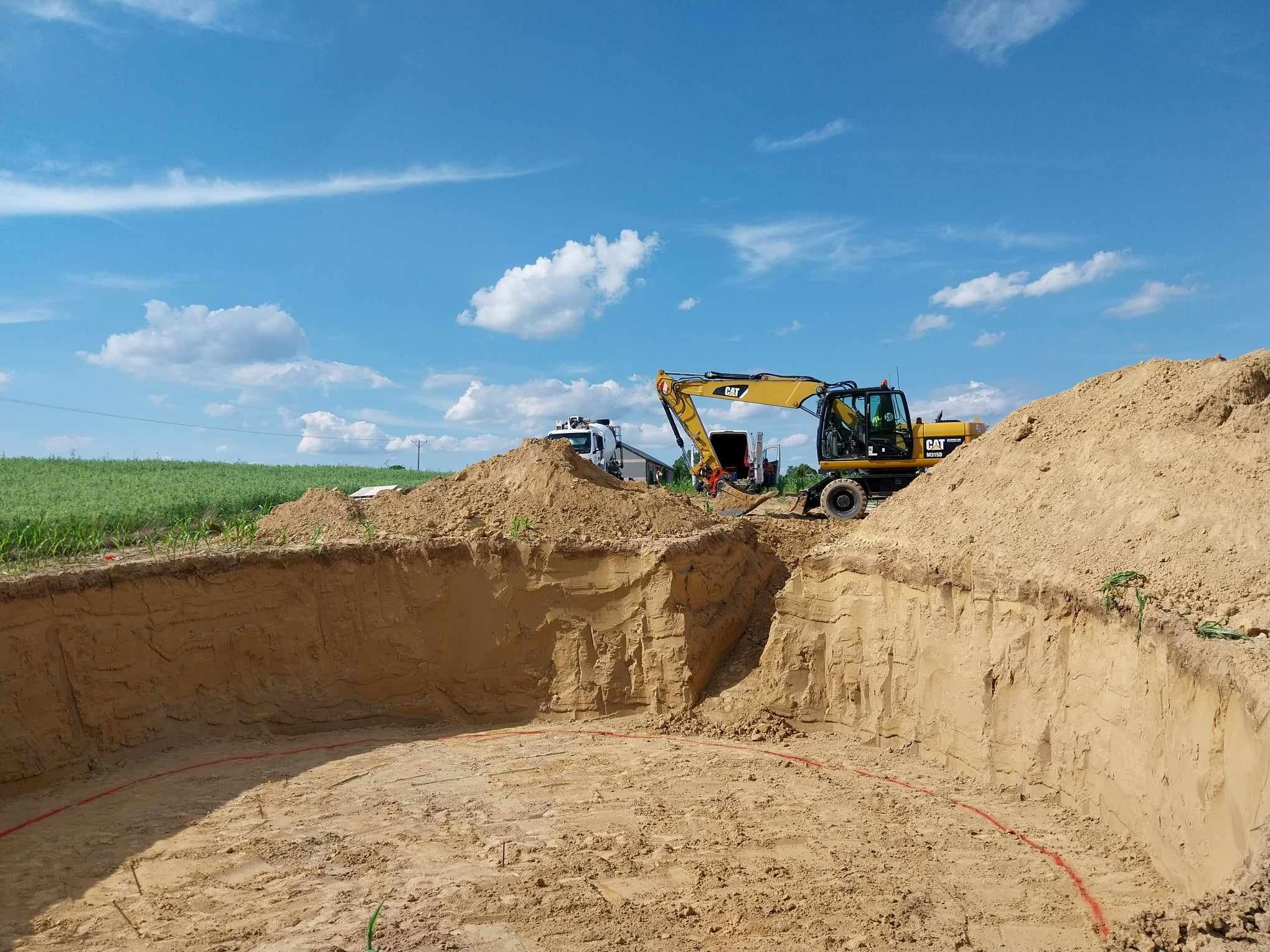 Żółta koparka CAT w trakcie robót ziemnych przy wykopie o okrągłym kształcie, oznaczonym czerwoną linią, na tle błękitnego nieba z chmurami.
