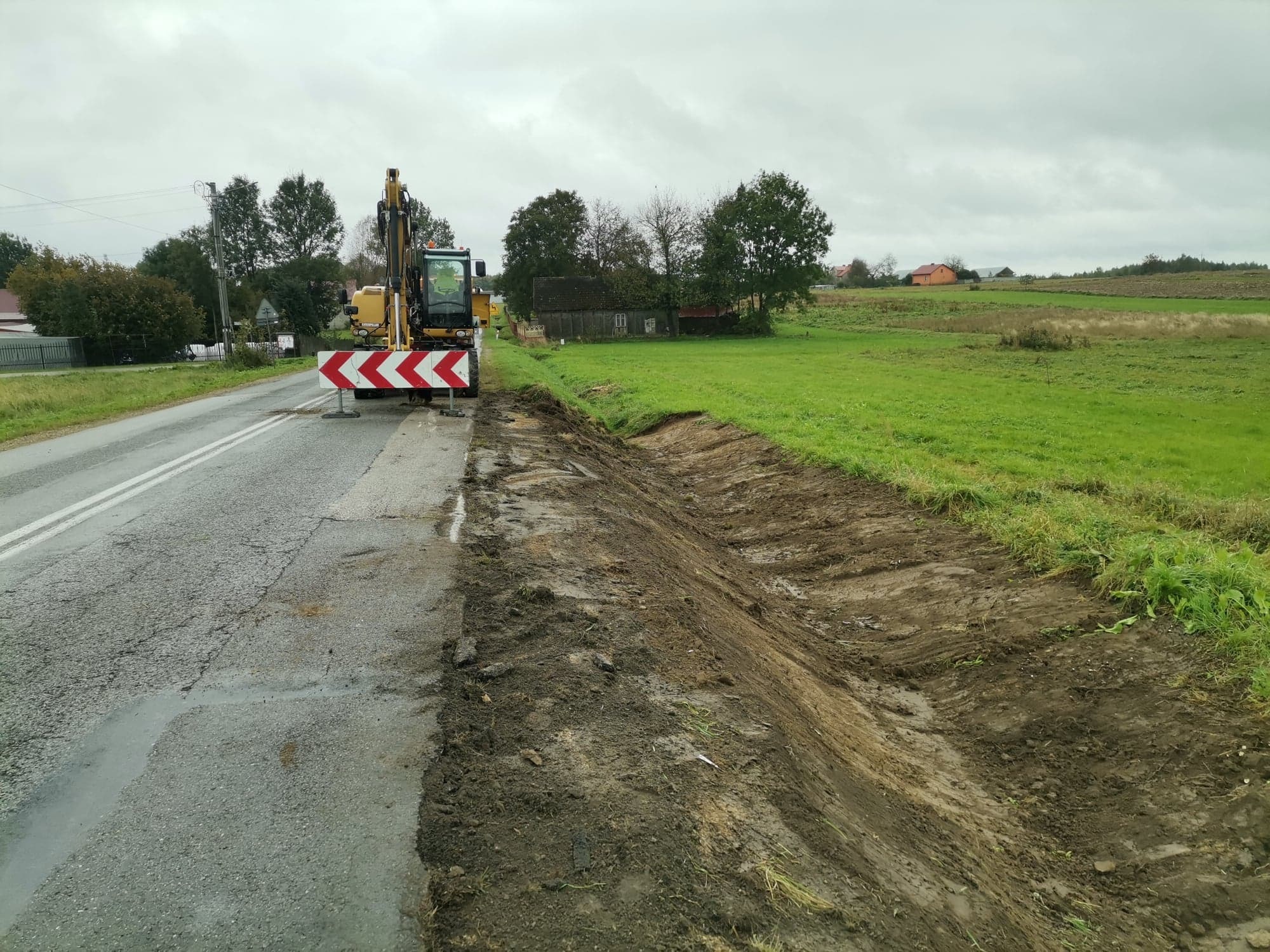 Żółta koparka przy drodze, obok wykopany rów w ziemi, widoczne znaki drogowe i zielone pole w tle, pochmurne niebo.