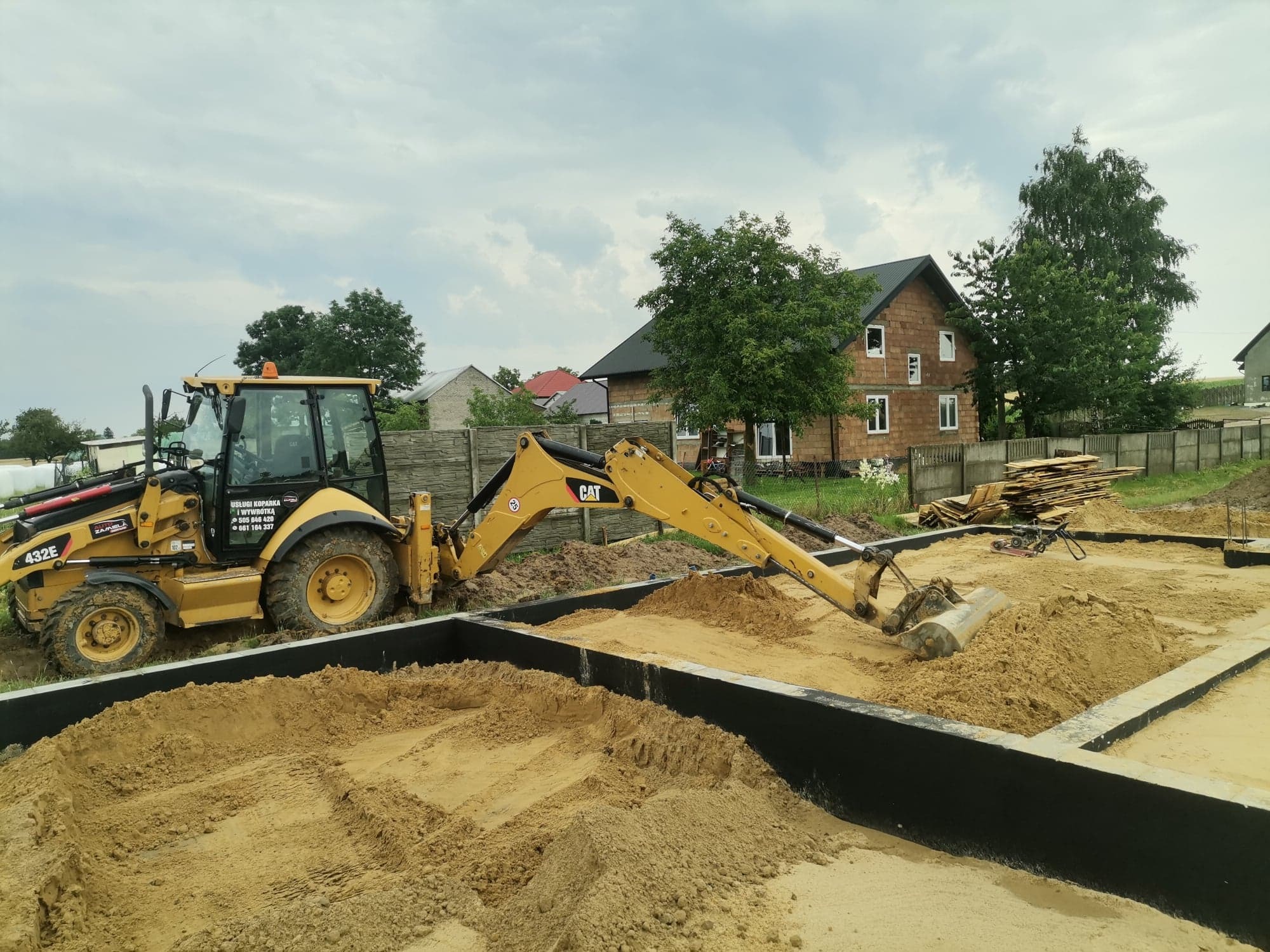 Żółta koparko-ładowarka CAT wypełnia piaskiem czarne szalunki fundamentowe na tle budowanego domu z cegły i pochmurnego nieba.