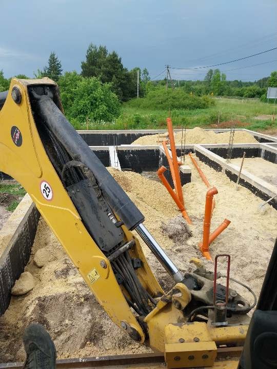 Żółta koparka w trakcie zasypywania piaskiem fundamentów, widoczne wystające rury kanalizacyjne, teren częściowo ogrodzony, w tle zieleń i słupy energetyczne.