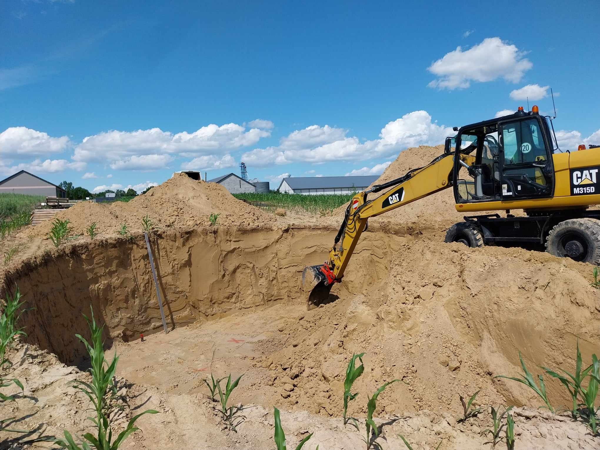 Żółta koparka CAT M315D w trakcie prac ziemnych, pogłębiania wykopu o okrągłym kształcie, widoczne ślady pomiarów na ziemi, w tle budynki gospodarcze i błękitne niebo z chmurami.
