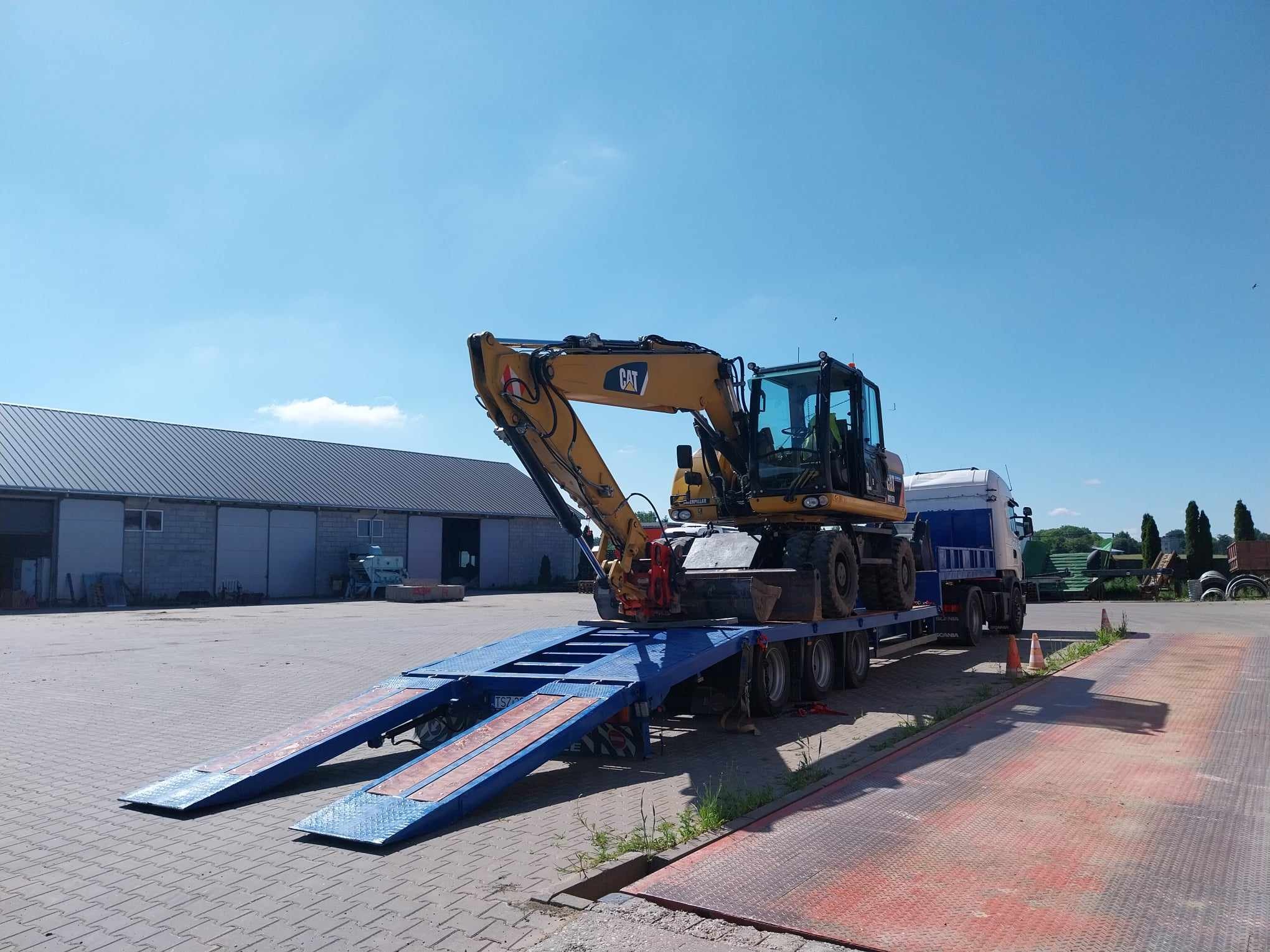Żółta koparka CAT na niskopodwoziowej naczepie z najazdami, transportowana przez białą ciężarówkę na tle szarego magazynu i błękitnego nieba.