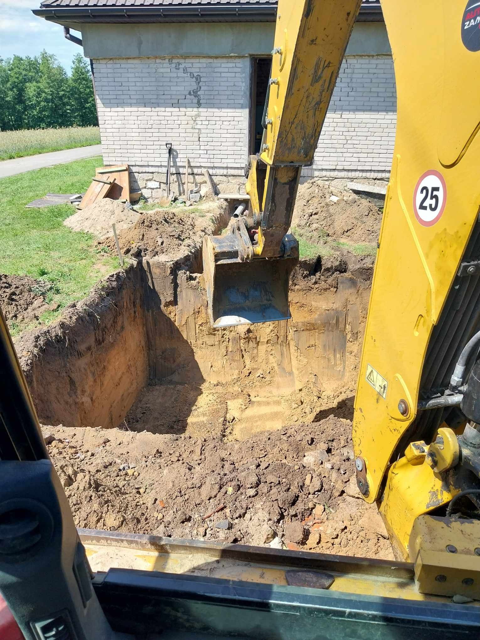 Żółta koparka z łyżką zagłębia się w ziemię, tworząc głęboki wykop, widok z kabiny operatora, w tle ceglany budynek i zielona trawa.