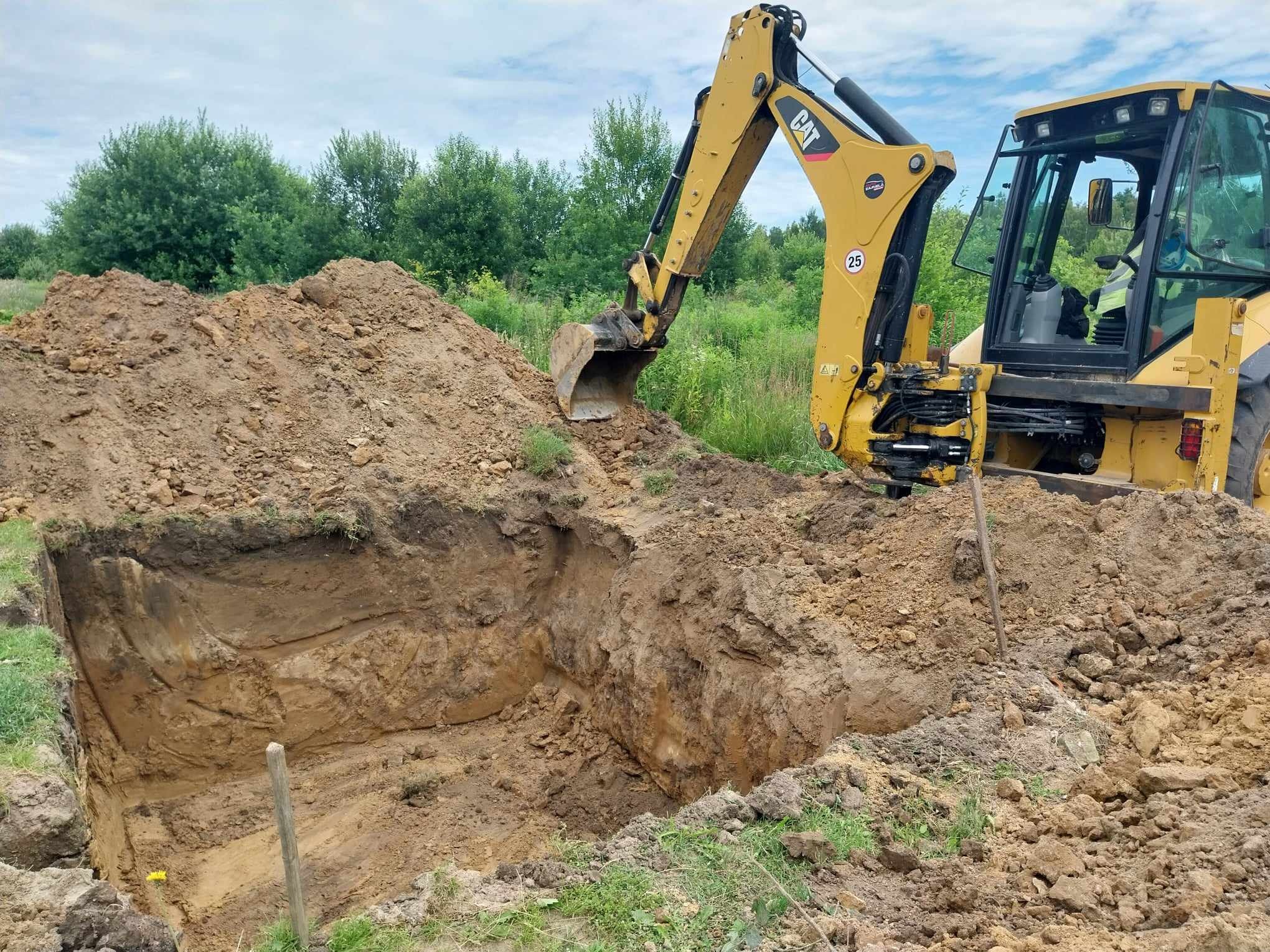 Żółta koparko-ładowarka CAT wykonuje wykop w ziemi, widoczne ślady pracy łyżki, w tle zieleń i drzewa, na pierwszym planie pryzma ziemi.