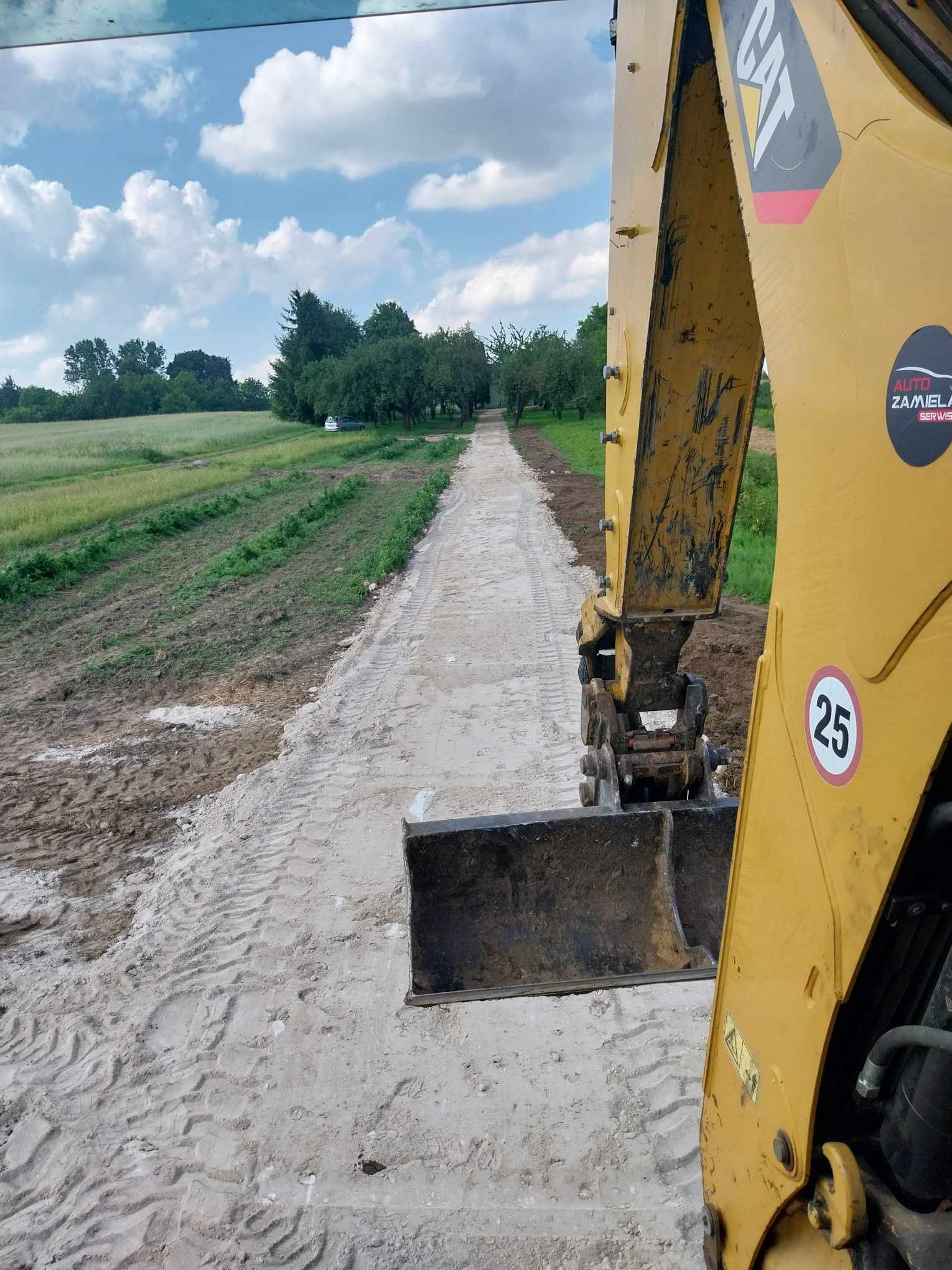 Żółta koparka CAT z łyżką na tle drogi gruntowej po świeżo wykonanych pracach, widoczne ślady opon i logo firmy na ramieniu maszyny.