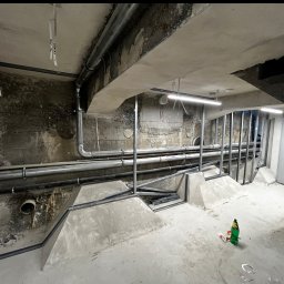 eR-Ka KAROL WOŹNICKI - Instalacja rur i konstrukcji metalowych w surowym wnętrzu remontowanego mieszkania. Widoczne elementy instalacji pod sufitem i na ścianach, oraz fragment schodów.