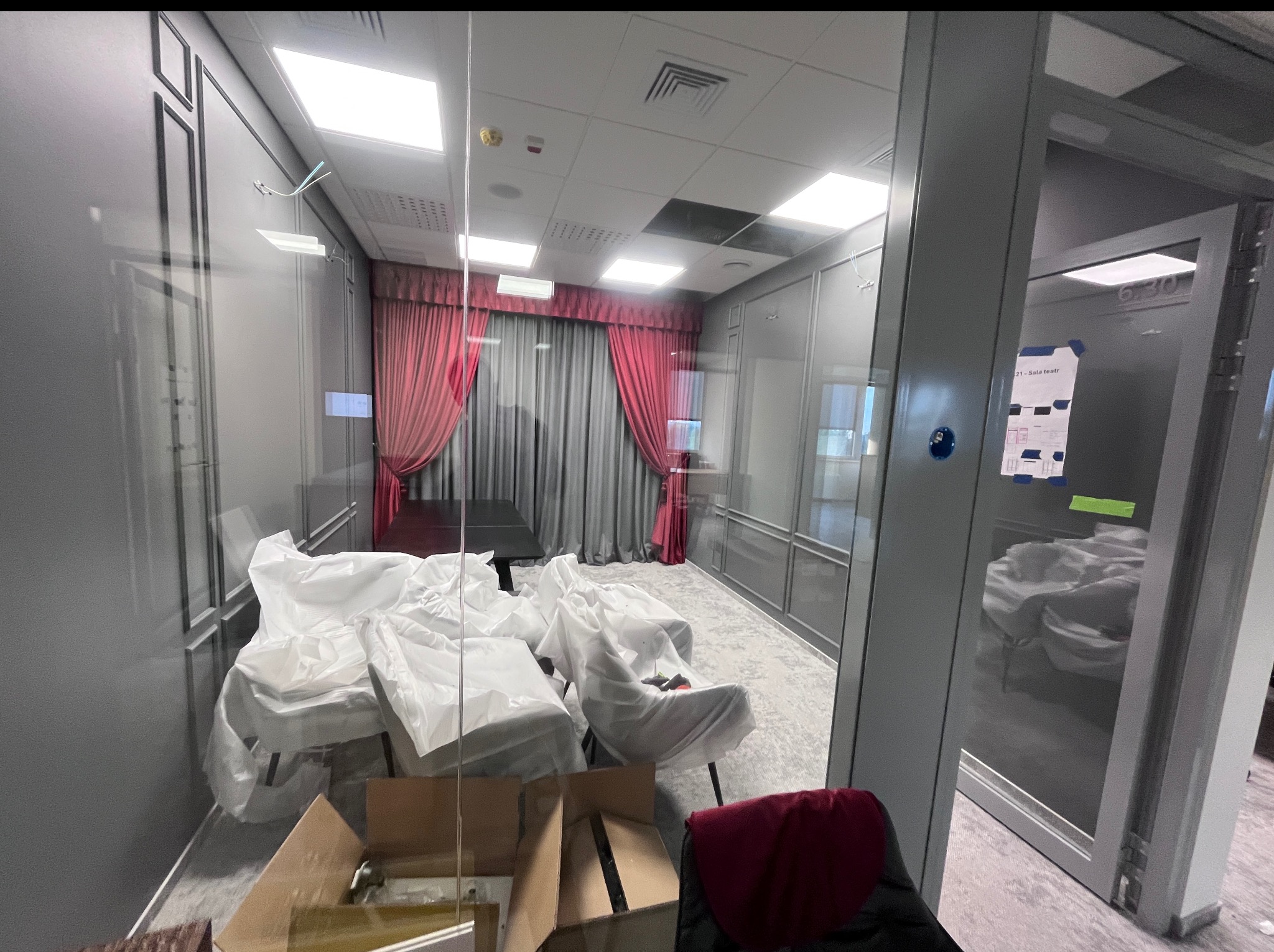 Pokój biurowy przed remontem: meble zakryte folią, kartony na podłodze, widoczne szare ściany i czerwone zasłony. Przygotowania do prac remontowych.