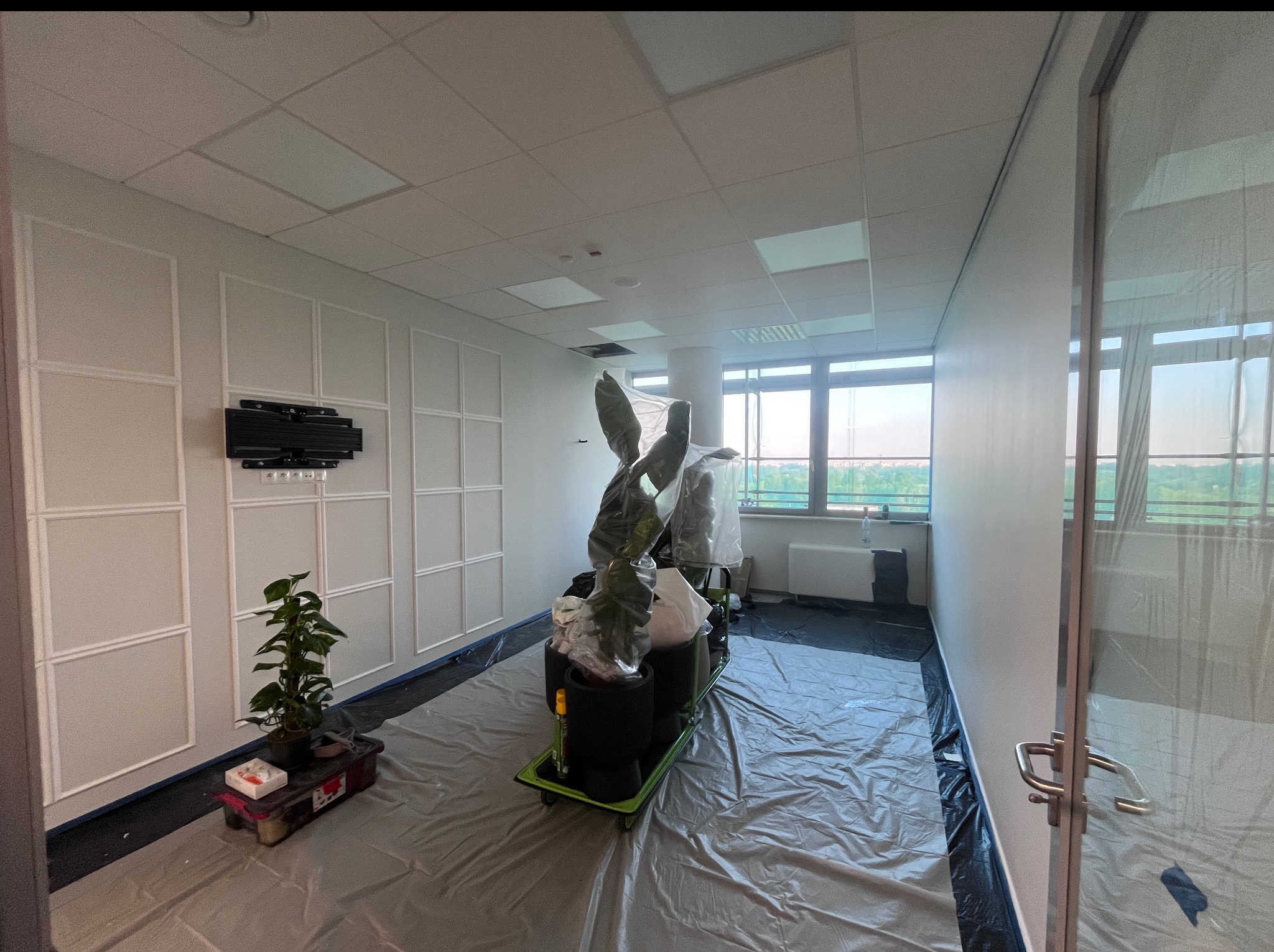 Przygotowanie biura do malowania: folia ochronna na podłodze, rośliny zabezpieczone folią, ściany z dekoracyjnymi panelami, widok na miasto z okna.