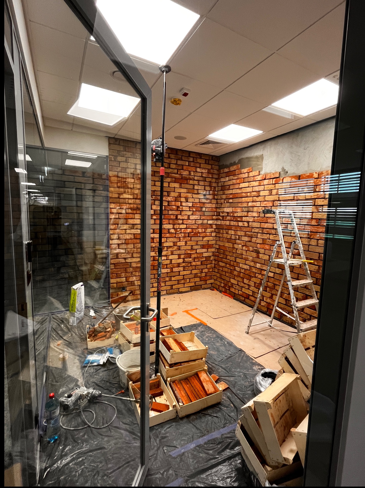 Wnętrze w trakcie prac z cegłą dekoracyjną na ścianie. Widoczna drabina, skrzynki z cegłami, narzędzia i folia ochronna. Częściowo wykończona ściana z cegły.