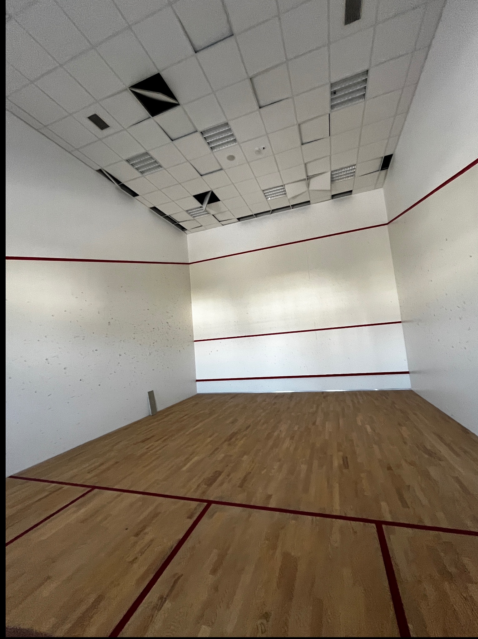 Wnętrze kortu do squasha z drewnianą podłogą, białymi ścianami z czerwonymi liniami i uszkodzonym sufitem podwieszanym. Widok z perspektywy gracza.